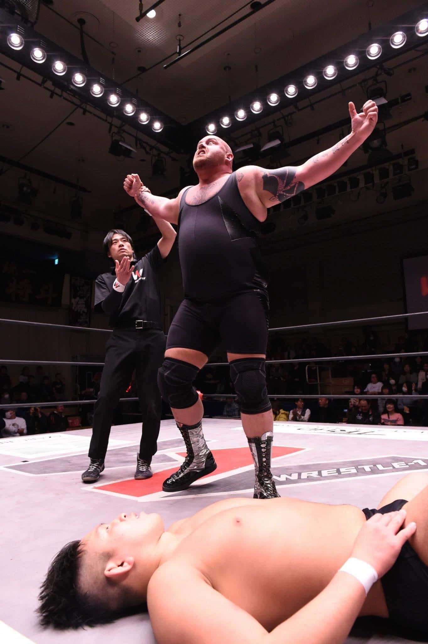Bam Bam Bigelow WWE 擊敗對手慶祝勝利 壁紙 — 一位肌肉發達的摔角手在燈光明亮的摔角場內，勝利地高舉雙臂，慶祝擊敗倒下的對手。