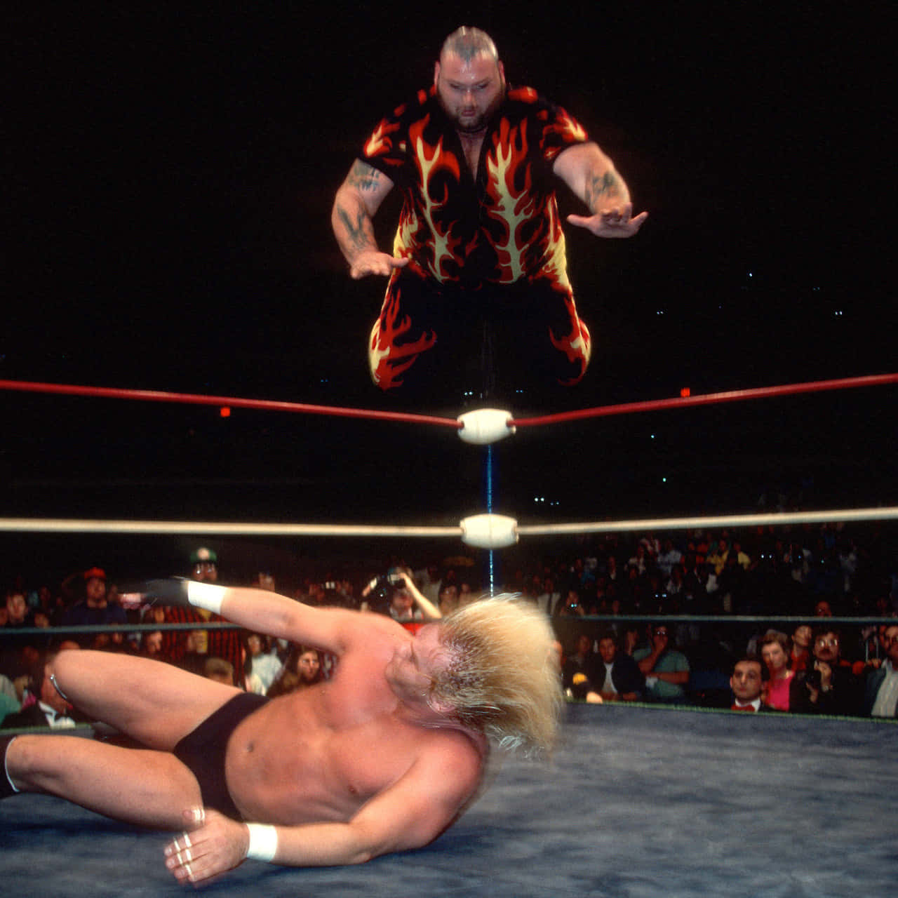 Bam Bam Bigelow 攻擊 Barry Windham 壁紙 — 一名身穿火焰圖案襯衫的摔角手從頂繩跳向躺在擂台上的對手。