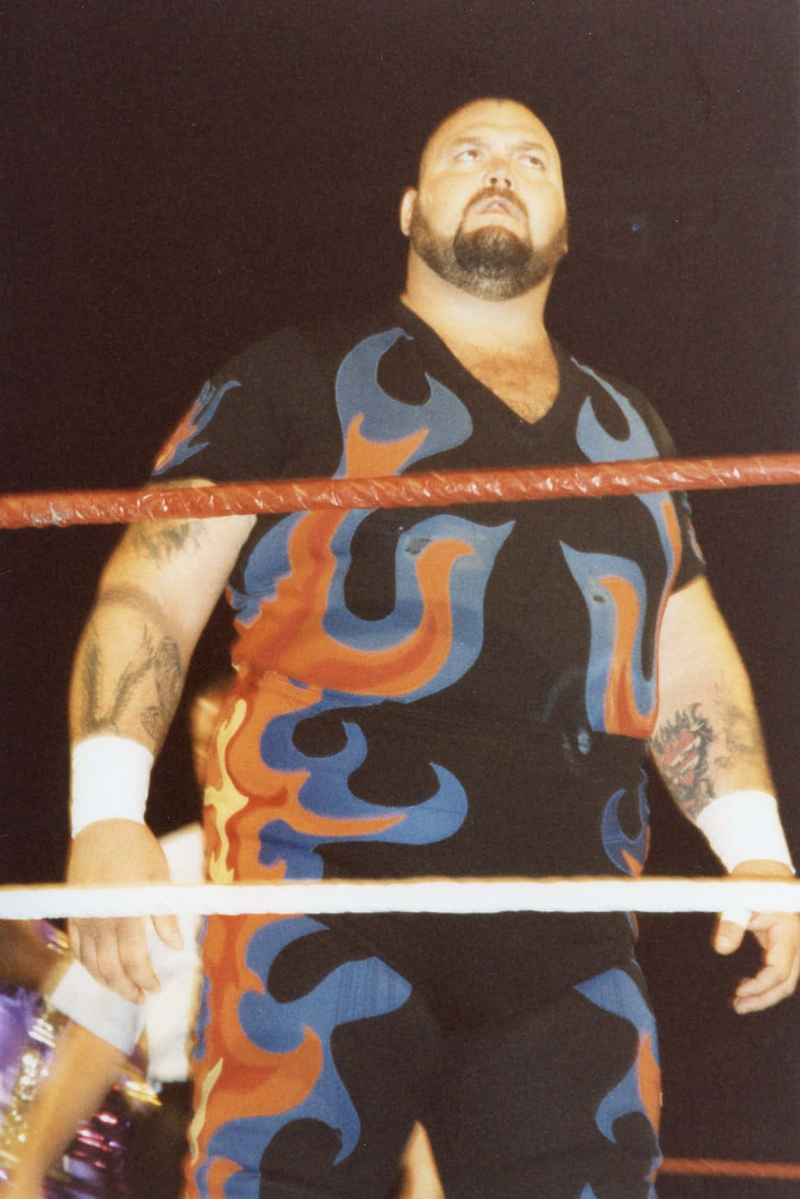 Bam Bam Bigelow 1994 香港溫布利體育館 壁紙 — 一位肌肉發達的摔角手穿著火焰圖案的黑色服裝，站在有紅色繩索的摔角擂台內，場館燈光璀璨。