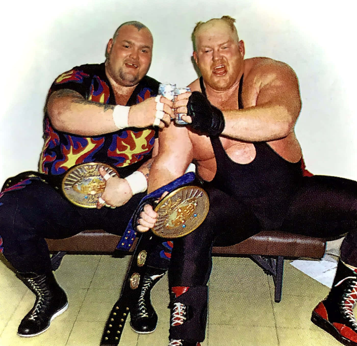 Bam Bam Bigelow 與 Big Van Vader 庆祝勝利 壁紙 — 兩名肌肉發達的摔角手身穿色彩繽紛的裝備，坐在長椅上，一邊拿著冠軍腰帶，一邊碰罐慶祝。