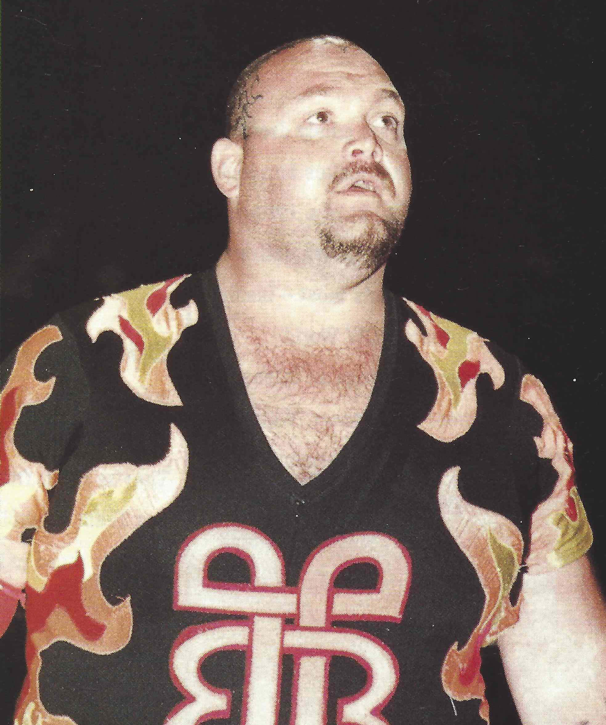 Bam Bam Bigelow WCW Magazine 1999 當代寫真 壁紙 — 一個光頭留山羊鬍的男人穿著黑色V領衫，胸前有火熱的橙黃紅火焰圖案和白色風格化標誌，背景為深色。