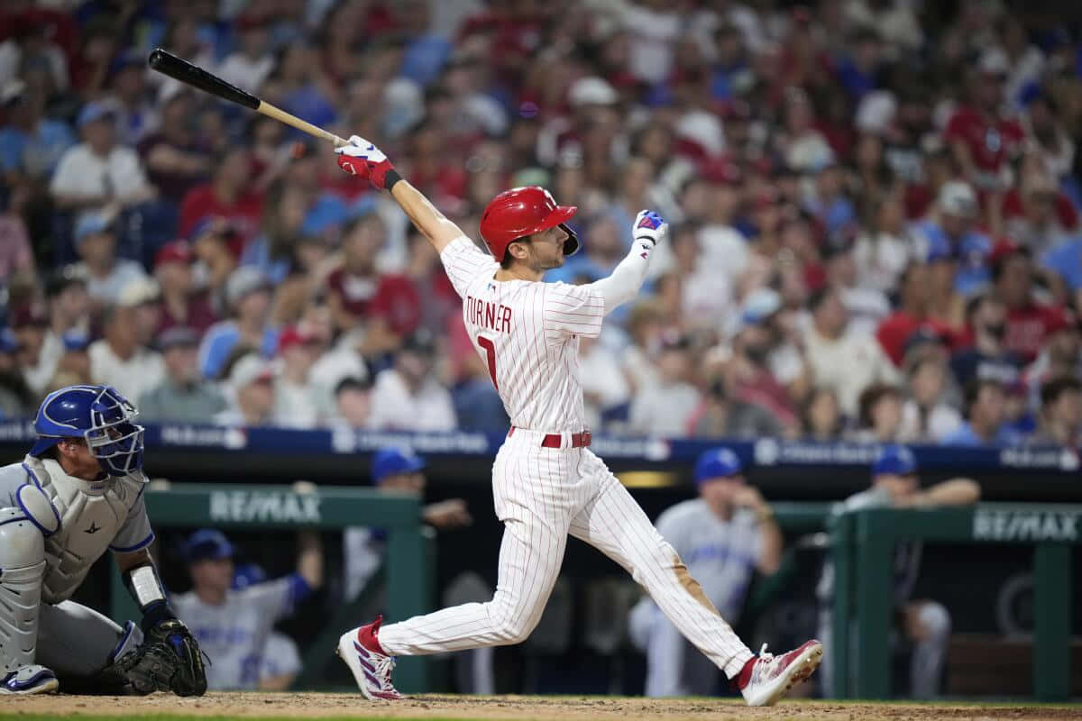 Trea Turner Phillies 桌布 壁紙 — 戴紅色頭盔、身穿白色細條紋球衣的棒球手在夜間比賽中強力揮棒，背景有觀眾歡呼。