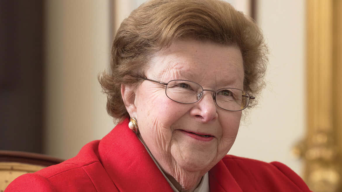 Barbara Mikulski 肖像照 壁紙 — 一位戴眼鏡、笑容溫暖的老年女性，在室內穿著一件鮮紅色的外套。