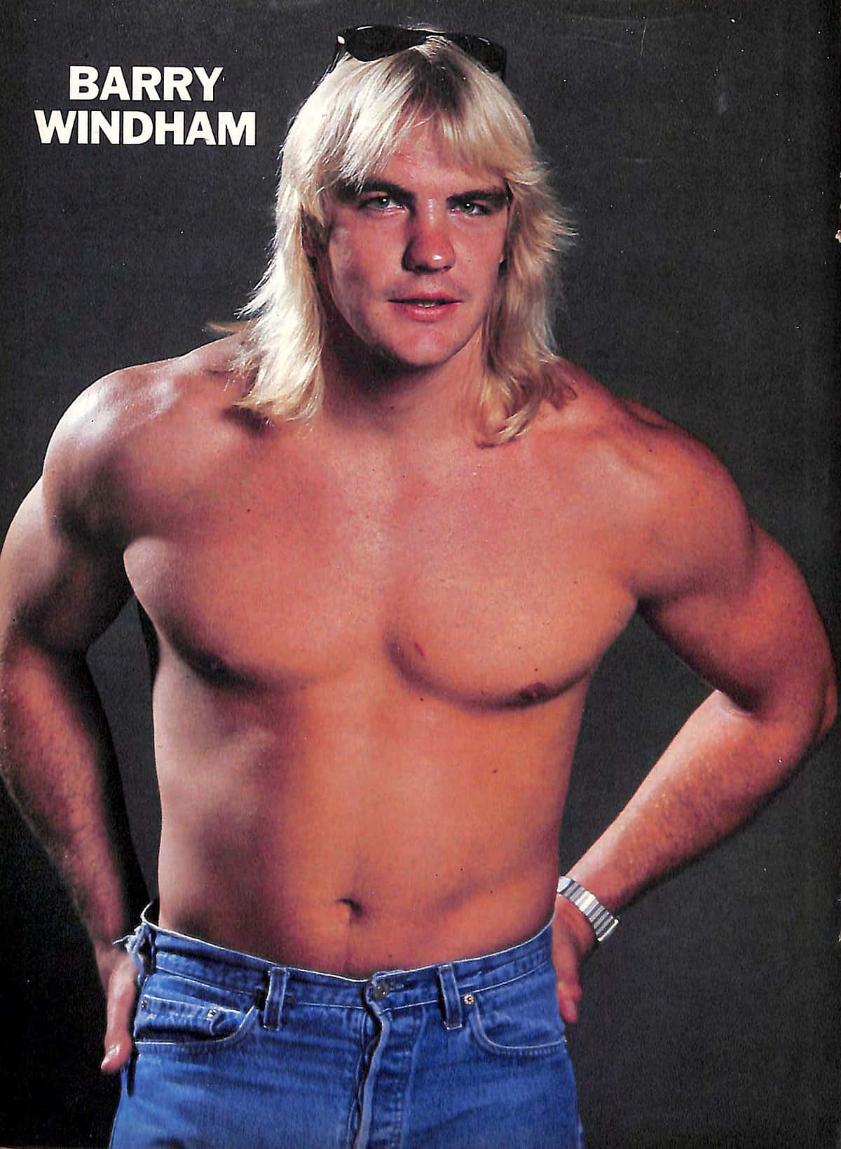 Barry Windham穿藍牛仔褲 壁紙 — 一個肌肉發達的男人，長長的金髮，頭上戴著太陽眼鏡，在深色背景前赤裸上身，穿著藍色牛仔褲擺姿勢。