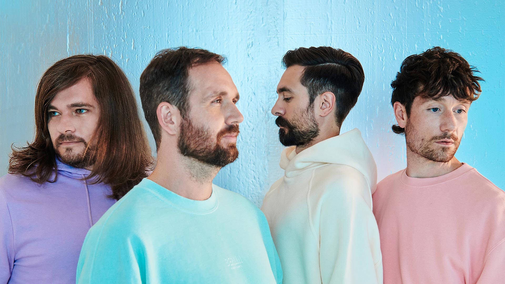 Bastille 淡彩攝影 壁紙 — 四個男人穿著粉彩色連帽衫和圓領衫，側身站在有紋理的藍色牆壁前，各自看向不同方向。