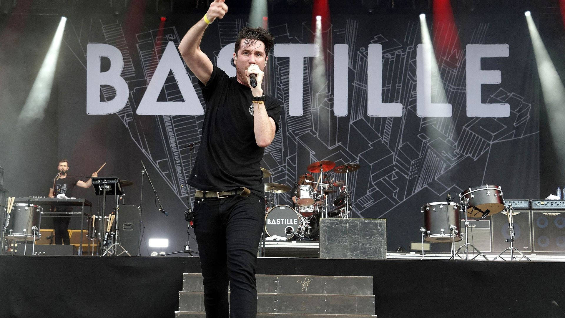 Bastille 演唱會現場演出 壁紙 — 一位身穿黑色衣服的男歌手在明亮的聚光燈下與樂隊在'BASTILLE'背景前充滿活力地表演。