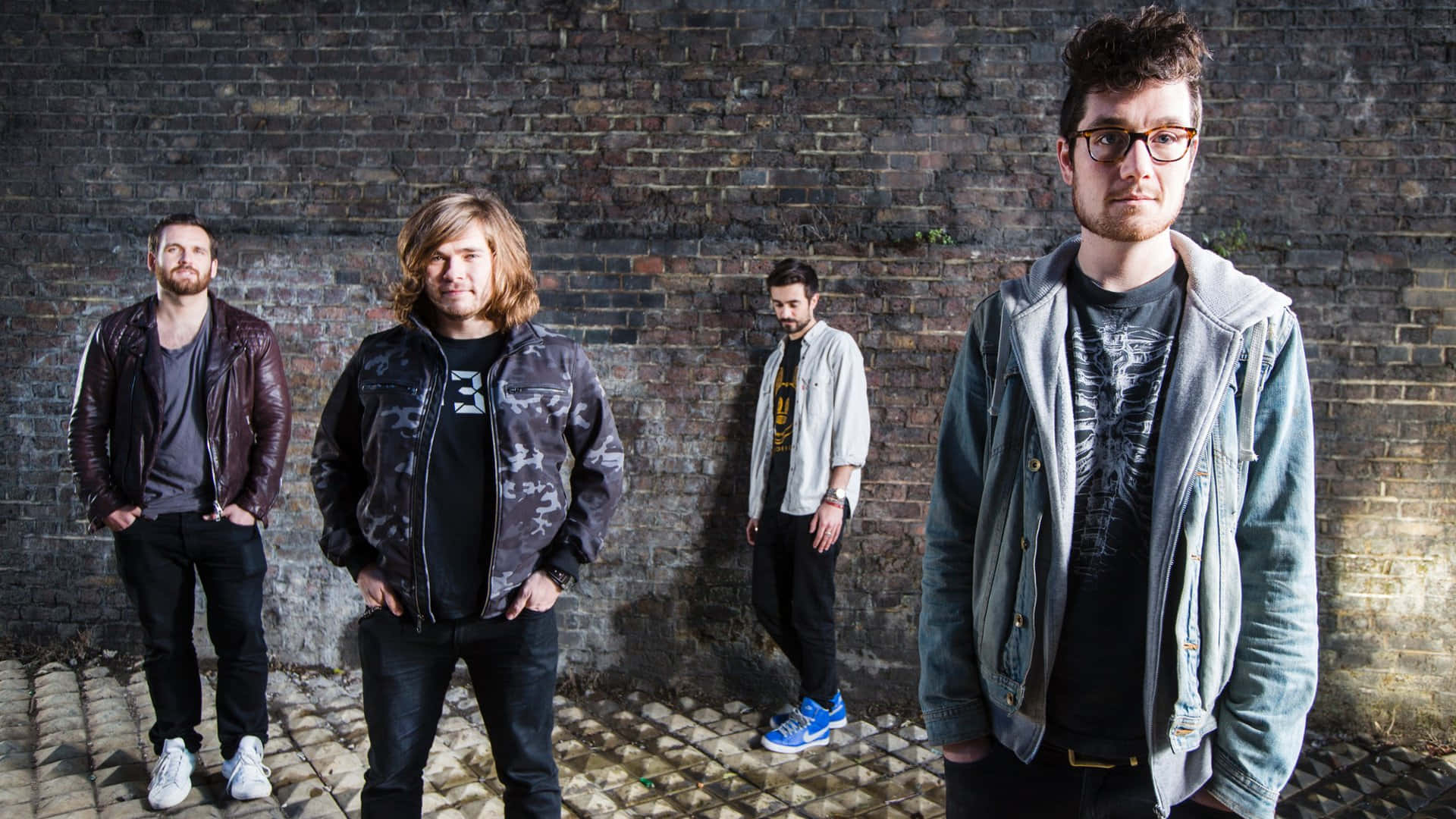 Bastille - 純粹歡樂 壁紙 — 四位身穿休閒街頭服飾的男士在明亮的日光下自信地站在風化的磚牆前。