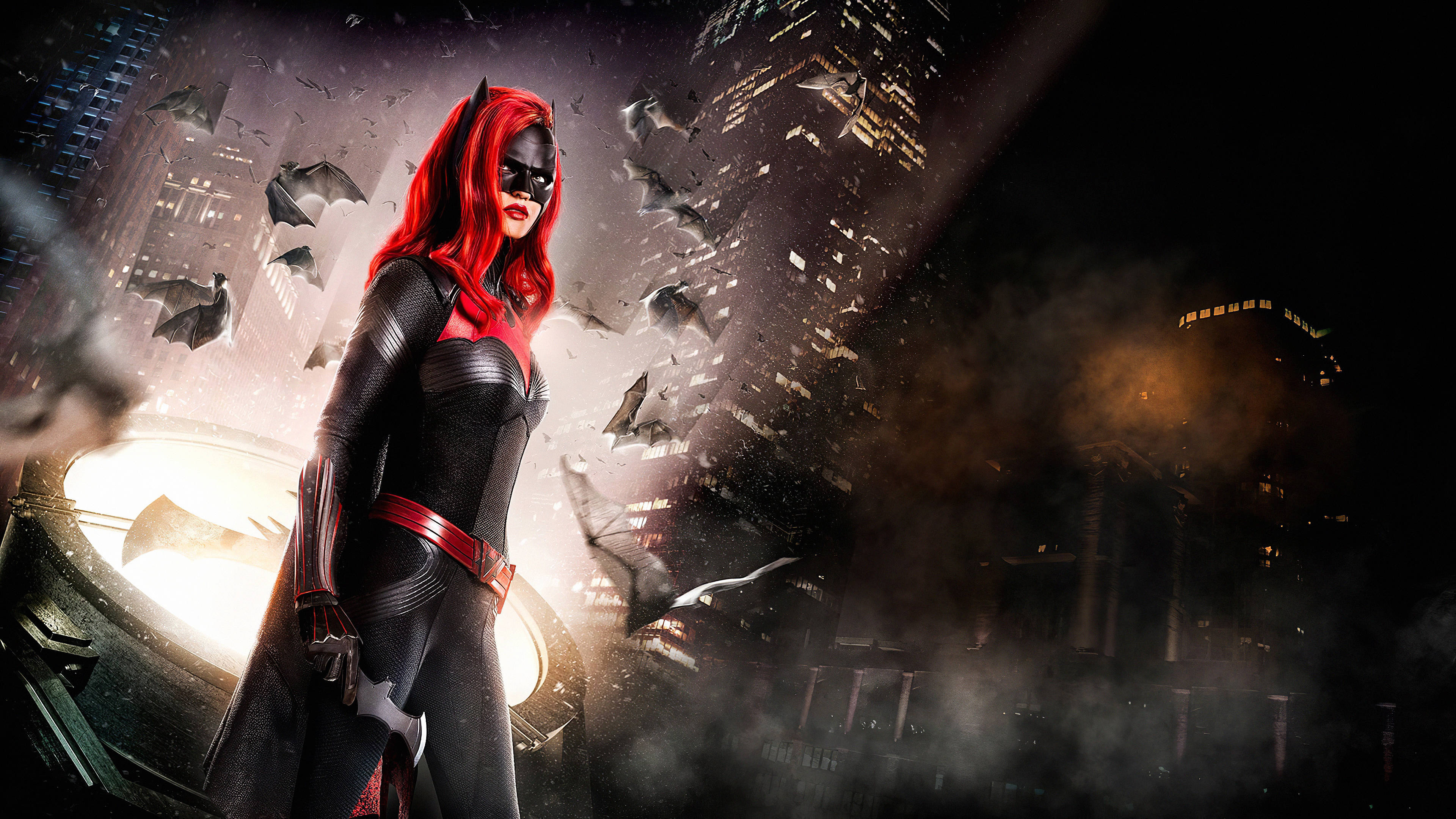 Batwoman Ruby Rose 夜間風采 壁紙 — 紅髮蝙蝠女俠在黑暗的城市景觀中英勇站立，蝙蝠在戲劇性的燈光下圍繞她飛舞。