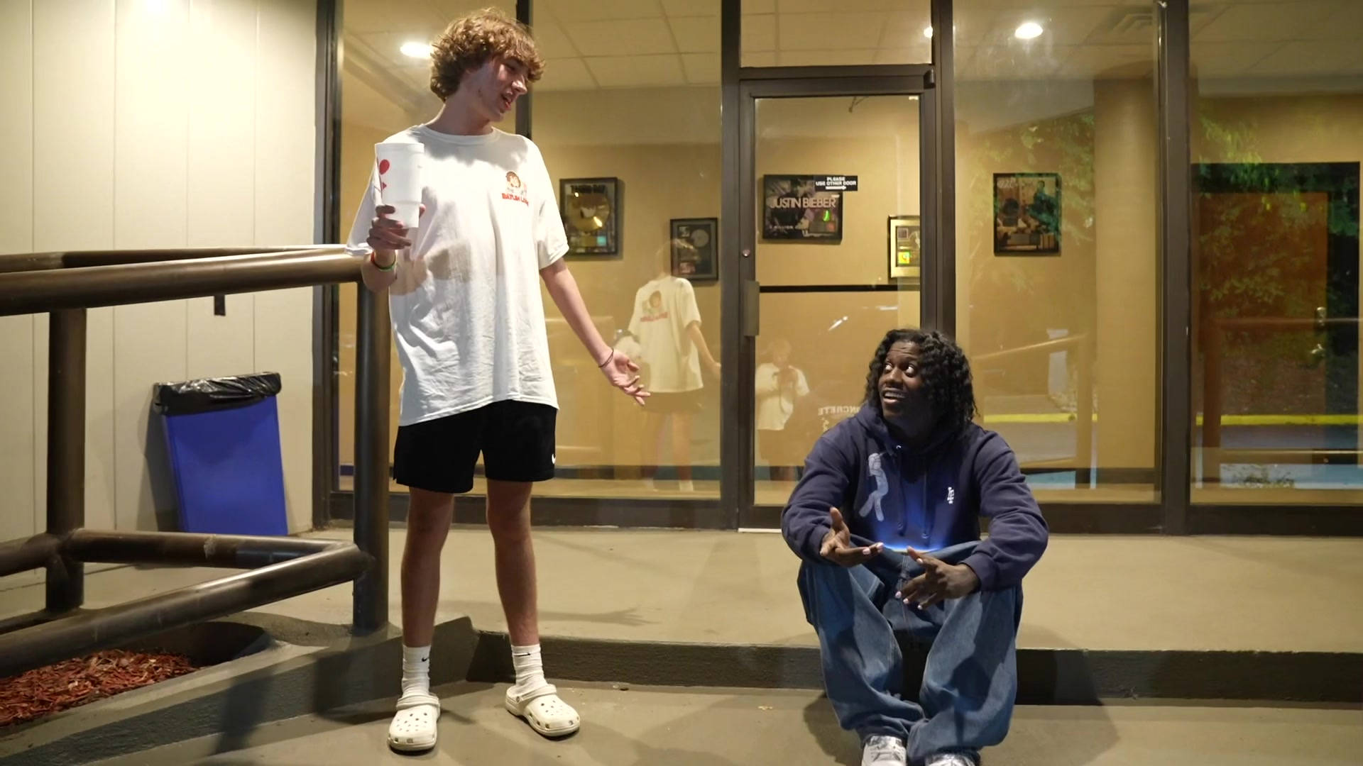 Baylen Levine 與饒舌歌手 Lil Yachty 壁紙 — 兩個年輕男子在夜晚的玻璃幕牆建築外大笑，一個站著拿著杯子，另一個生動地蹲著。
