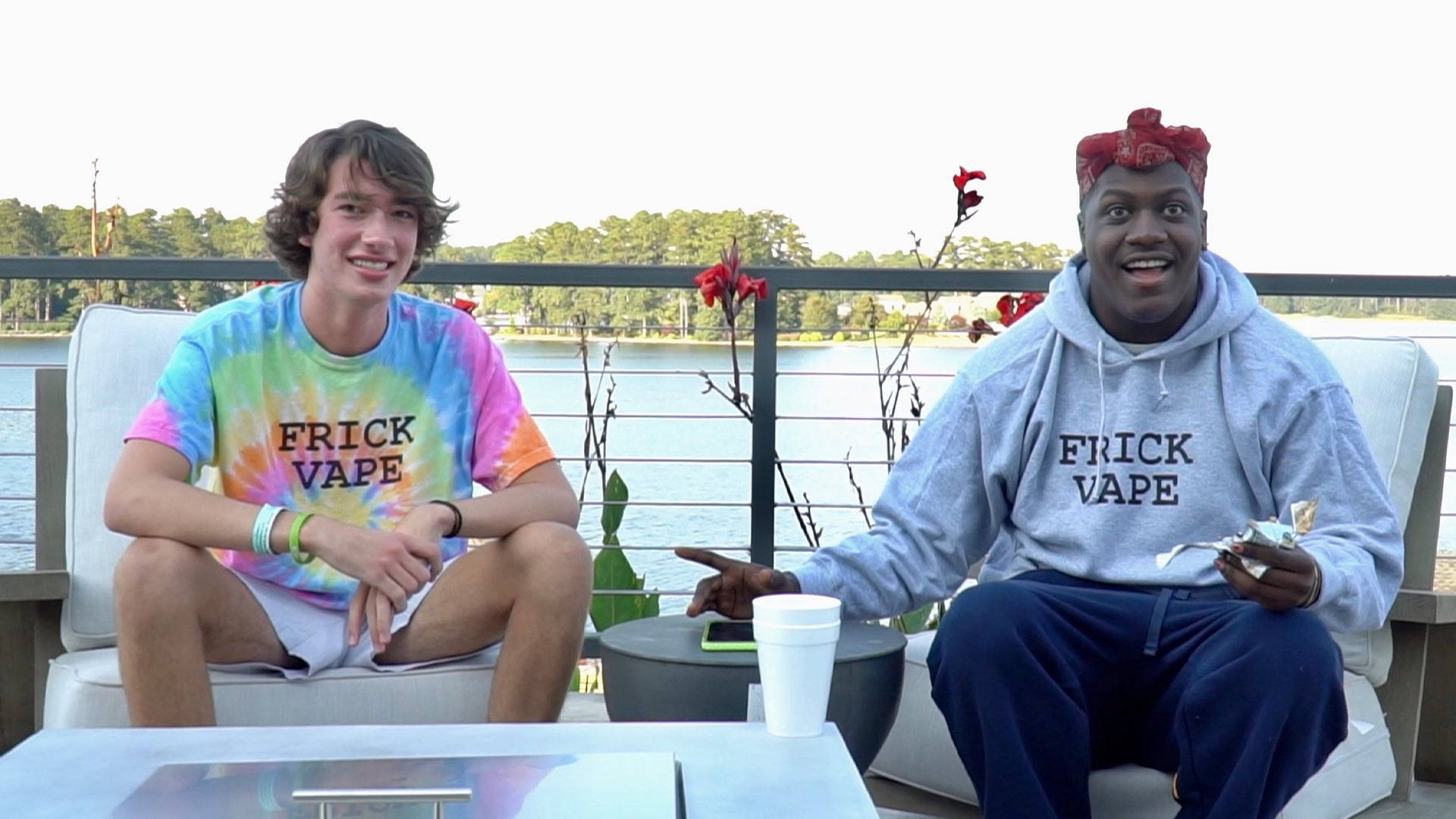 Baylen Levine 與饒舌歌手 Lil Yachty 壁紙 — 兩名微笑的年輕男子穿著相配的「FRICK VAPE」T恤，坐在陽台上，俯瞰著平靜的湖泊和紅花。