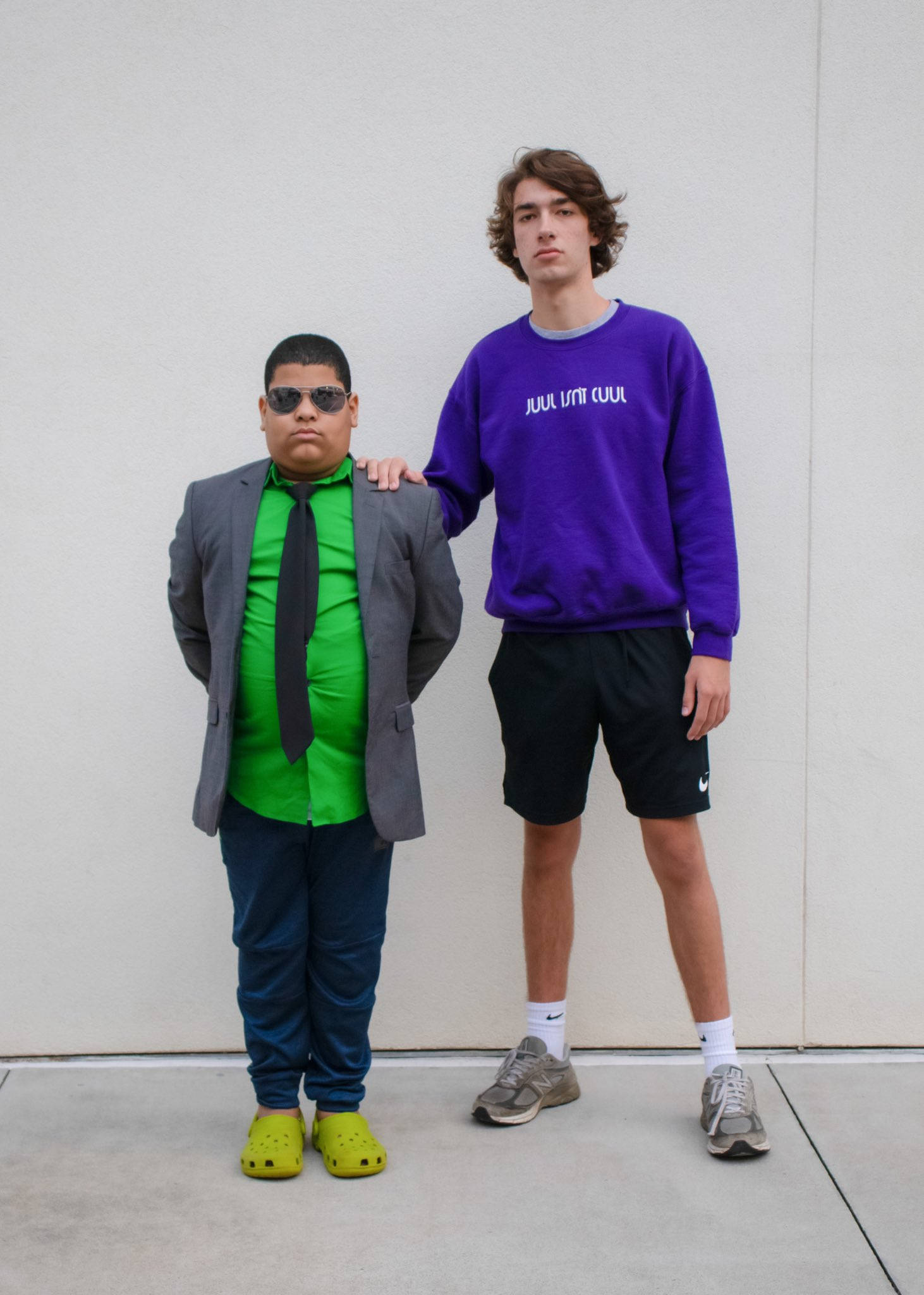 Baylen Levine 與 Super Sebas 壁紙 — 兩名年輕男子在素牆前擺姿勢，一人身穿綠色襯衫、灰色西裝外套和黃色crocs，另一人身穿紫色運動衫和黑色短褲。