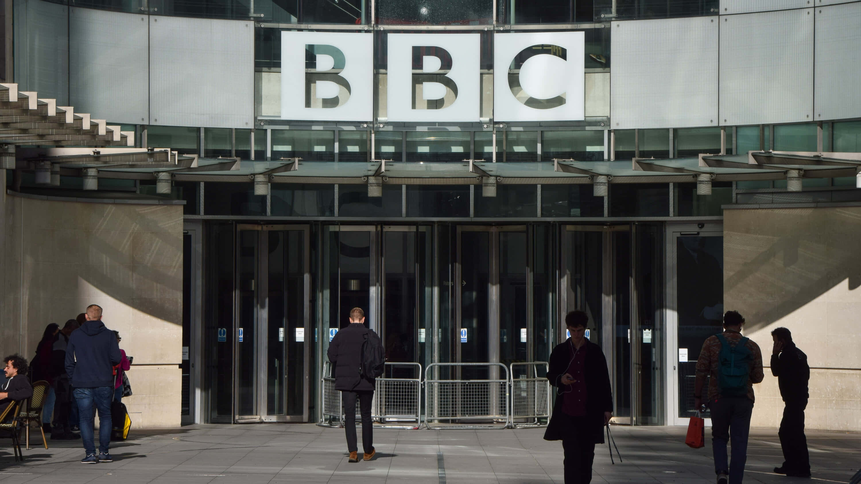 BBC大樓與少數人群圖 壁紙 — 人們在明亮的日光下走過BBC大樓的現代玻璃外牆。