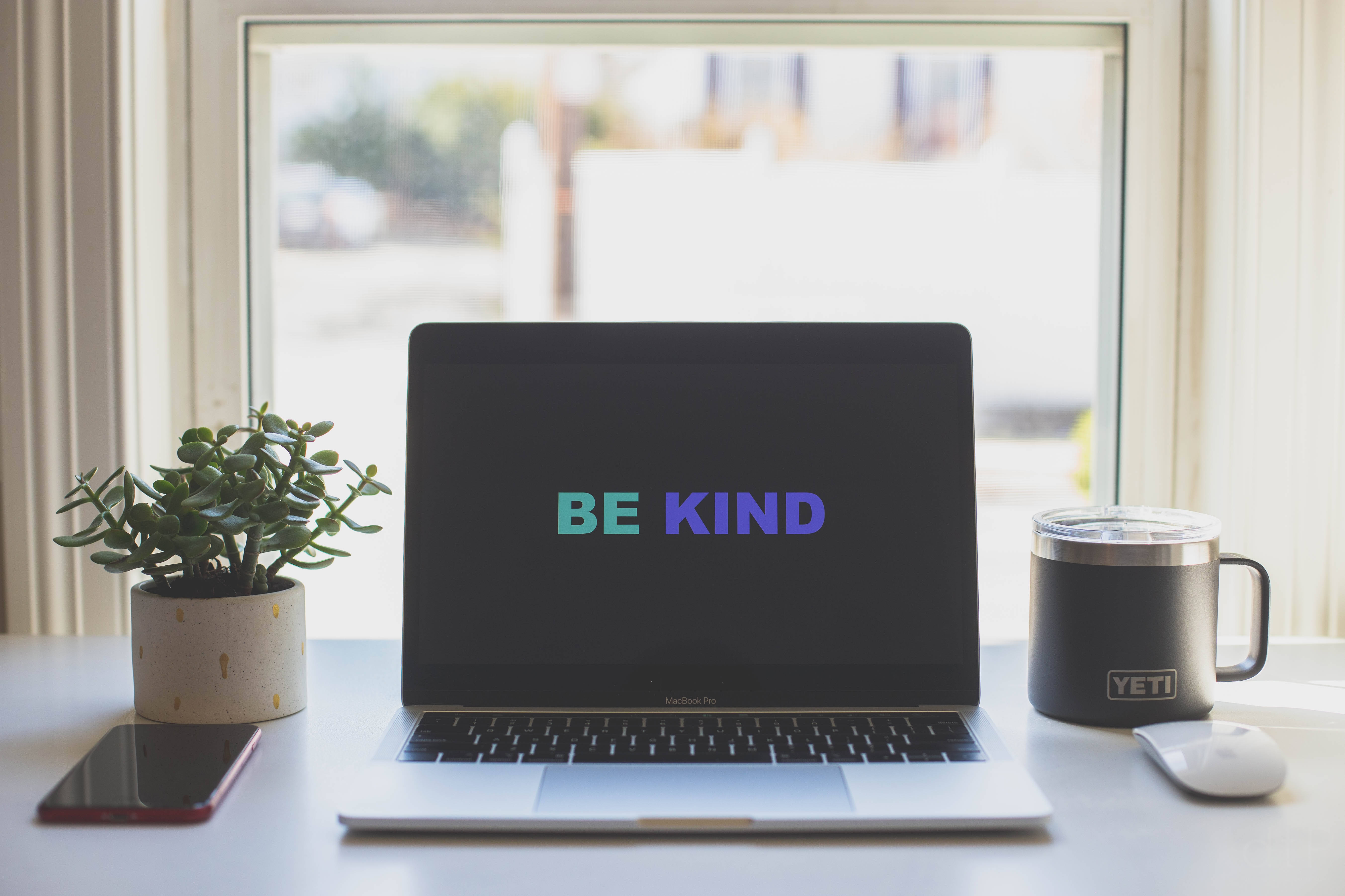 螢幕上嘅「Be Kind」勵志金句 壁紙 — 一部顯示「BE KIND」的筆記型電腦放在明亮的窗台上，旁邊有一棵植物、一個Yeti杯子和一部電話。