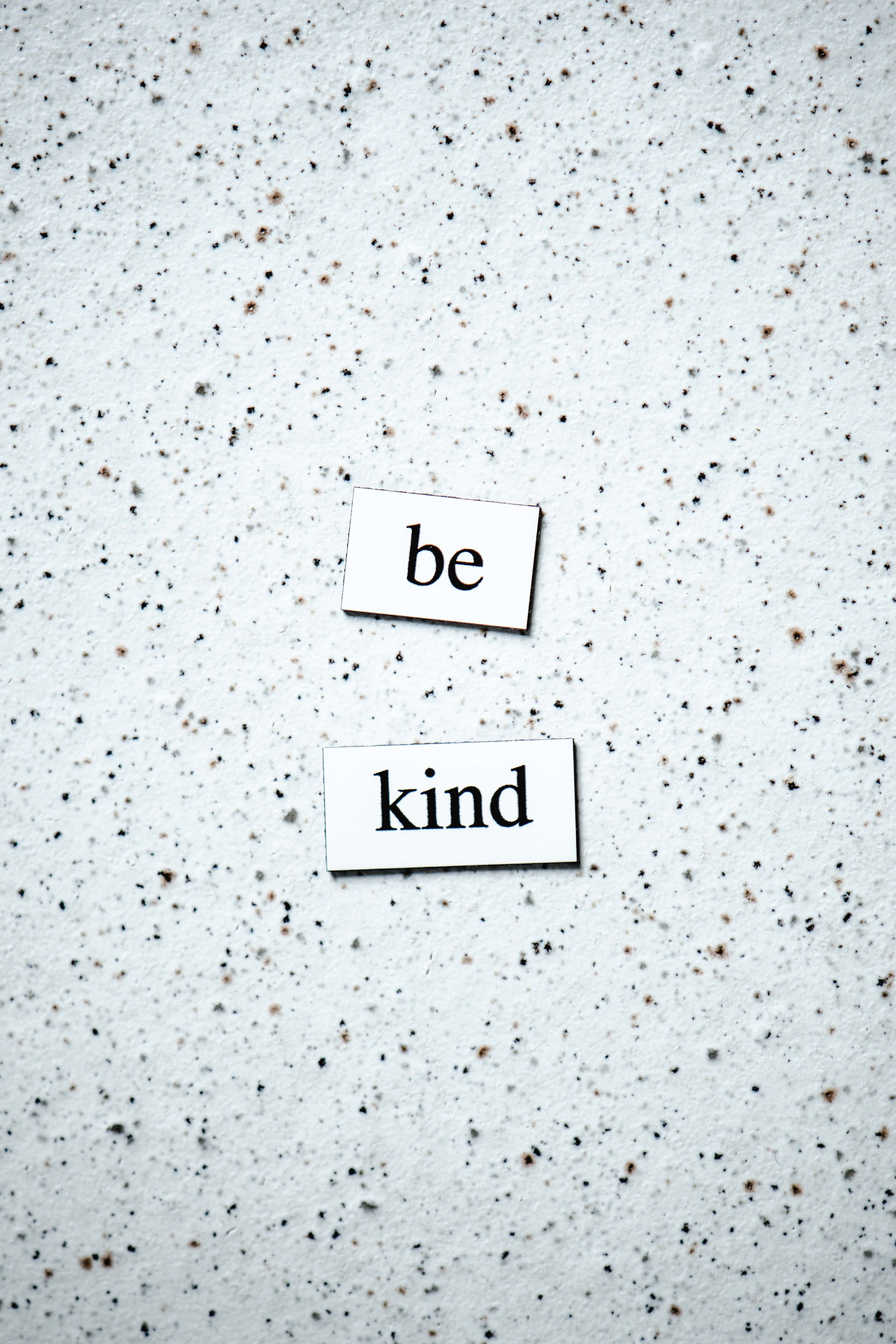 紙條上寫著「Be Kind」 壁紙 — 兩張白色卡片上印有黑色文字「be」和「kind」，放在斑駁的灰色混凝土表面上。