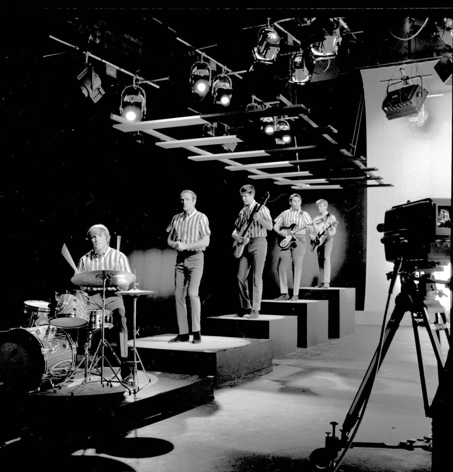 Beach Boys 1964年CBS電視節目拍攝 壁紙 — 一張1960年代樂隊在分層舞台上、在攝影棚燈光下、相機放在三腳架上表演的黑白照片。