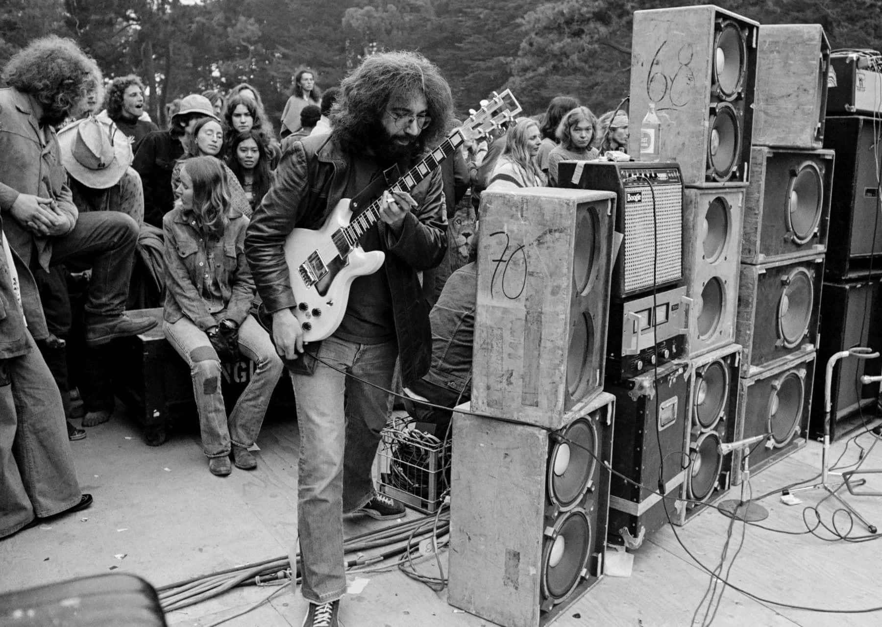  壁紙 — 一位留鬍鬚的結他手在1970年代戶外音樂會的舞台上，被堆疊的擴音器和人群包圍，彈奏白色電結他。