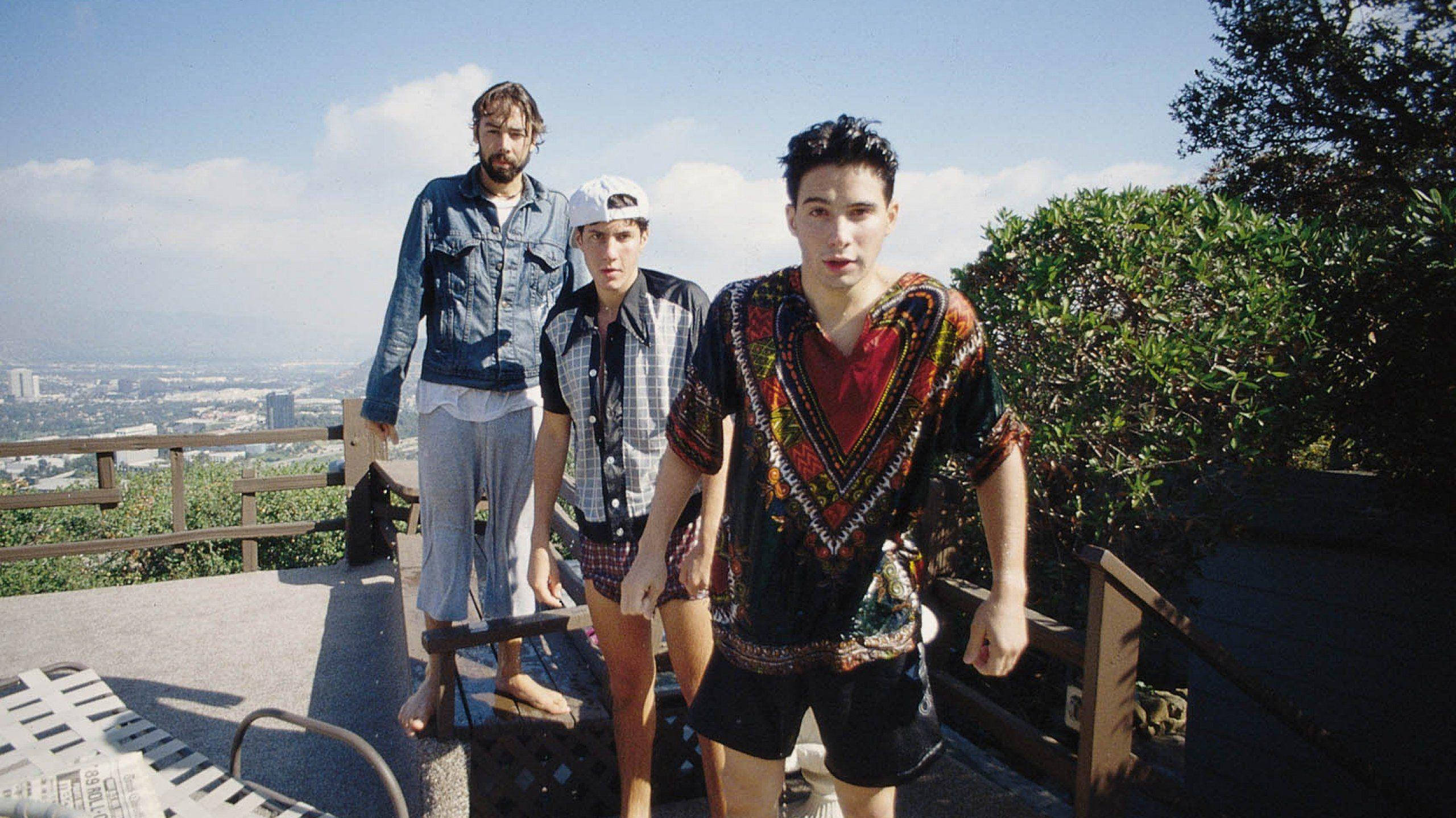 Beastie Boys《Hot Sauce Committee》專輯封面壁紙 壁紙 — 三名年輕男子穿著休閒夏季服裝，站在山坡上，俯瞰著晴朗藍天下的一座廣闊城市。