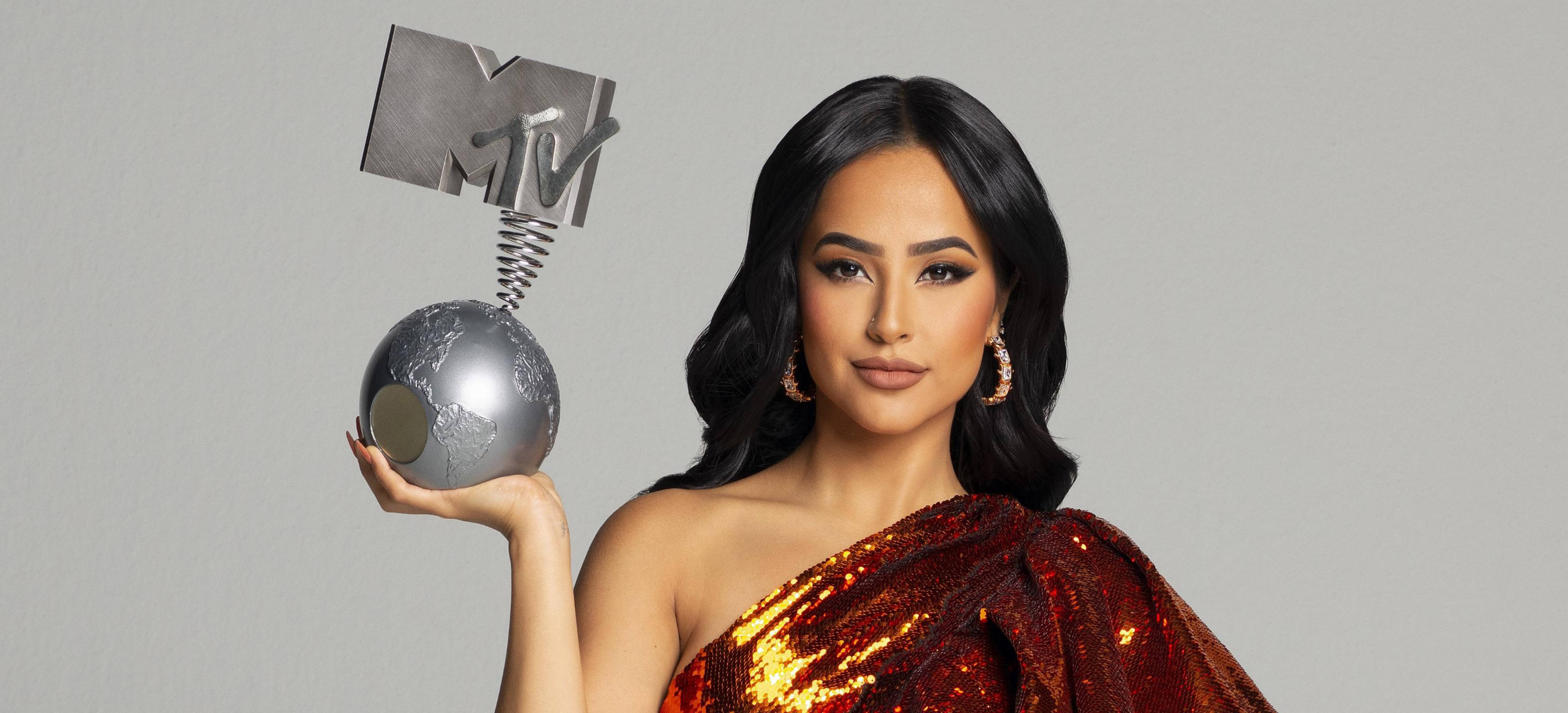 Becky G 2019 MTV 美洲音樂獎寫真 壁紙 — 一位身穿閃亮紅色長裙的女性，在純灰色背景前手持金屬質感的MTV Moon Person獎項。
