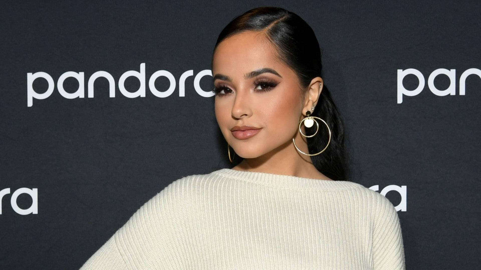 Becky G 出席 Pandora 活動 壁紙 — 一位深色頭髮、戴著金色圓環耳環的女性，穿著奶油色羅紋毛衣，在黑色Pandora品牌背景前擺姿勢。
