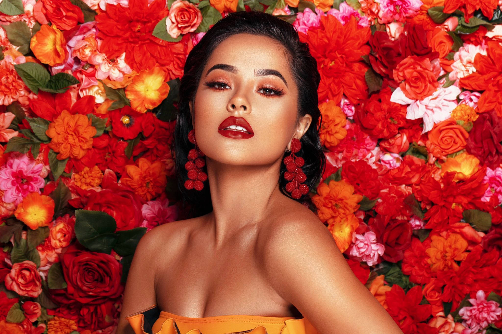 Becky G 為 ColourPop 品牌拍攝 壁紙 — 一位擁有鮮豔紅唇和相配耳環的女性，自信地站在由紅、橙、粉紅花朵組成的鮮豔花牆前。