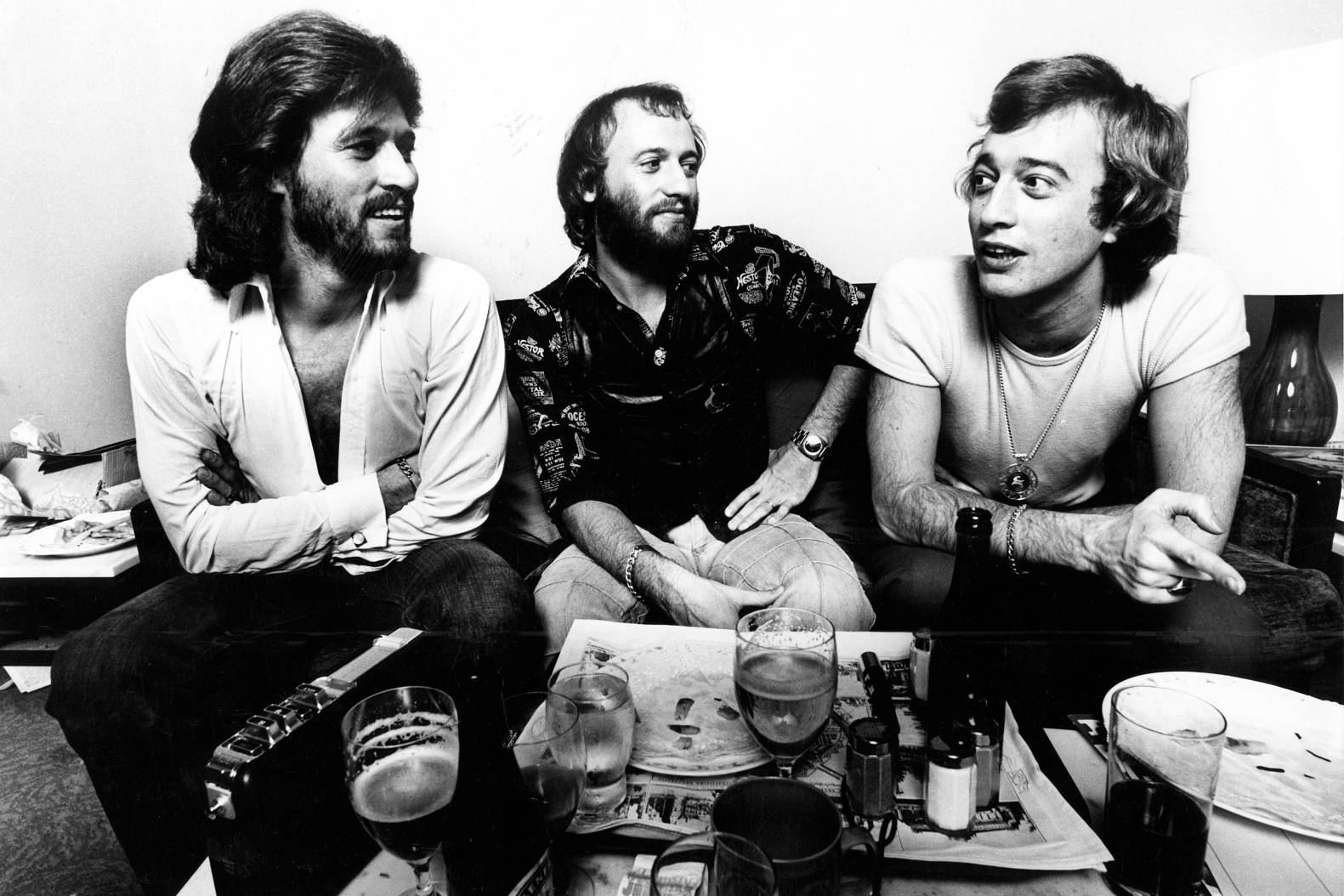 Bee Gees 1975 年阿姆斯特丹現場演出 壁紙 — 三位留鬍子、70年代髮型的男士休閒地坐在沙發上，周圍放滿飲品和雜亂的餐具。