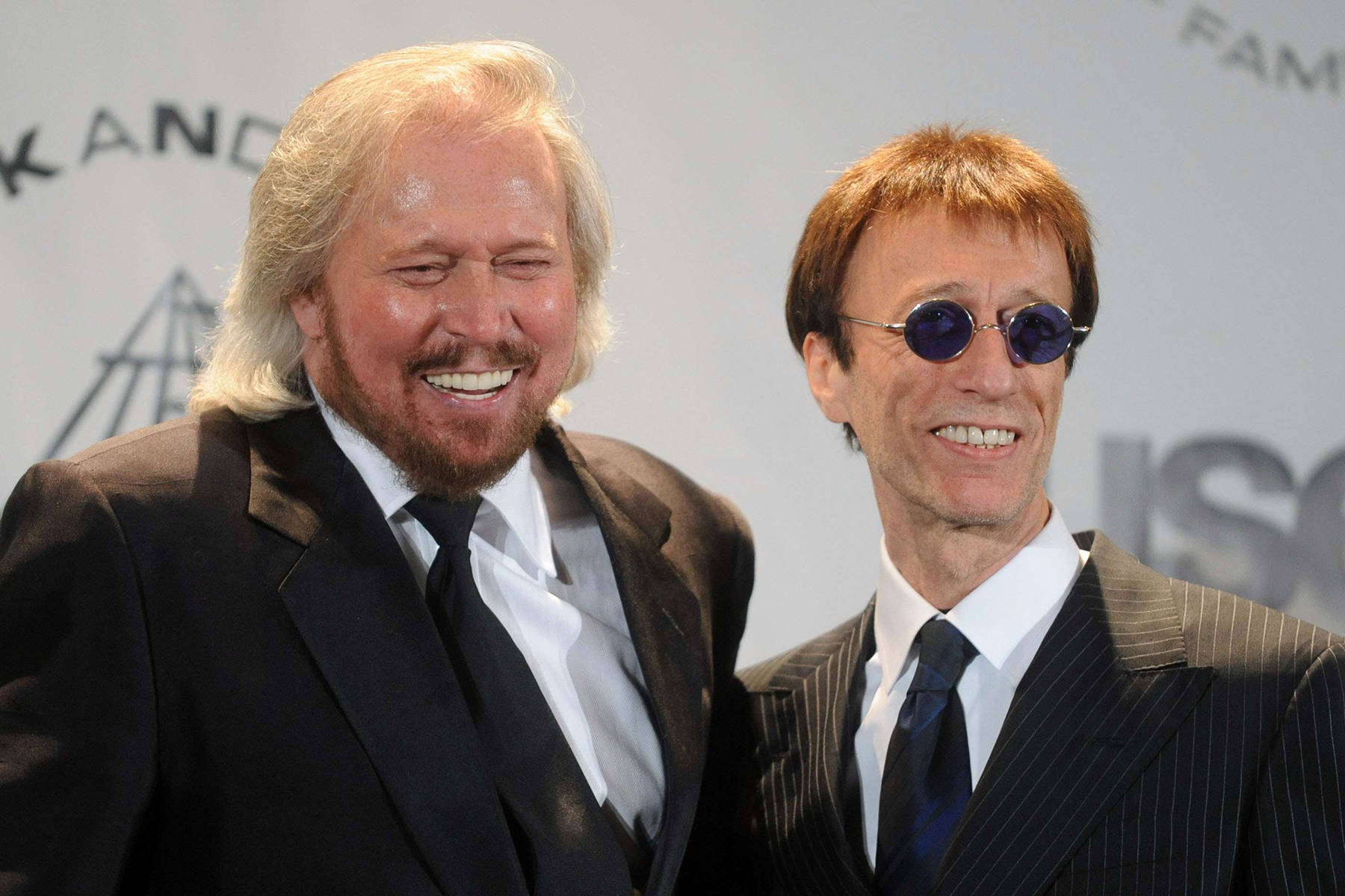 Bee Gees 巴里與羅賓·吉布 2010 年搖滾名人堂入選典禮 壁紙 — 兩位微笑的西裝男子，一位留著金色長髮，另一位戴著圓形太陽眼鏡，在活動中擺姿勢。