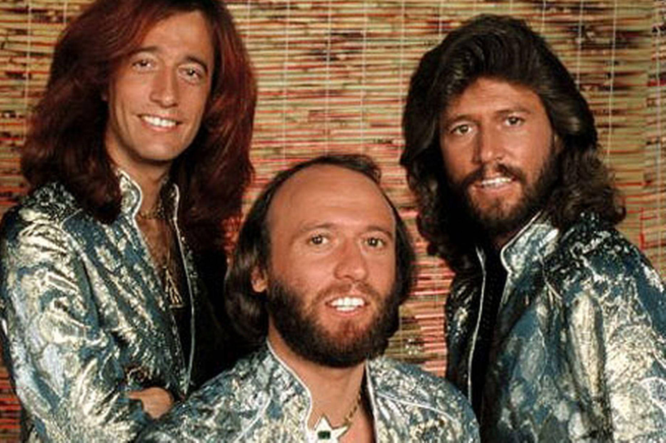 Bee Gees 澳洲流行樂團宣傳照 壁紙 — 三位笑容滿面、長髮、身穿閃亮銀色外套的男士，在編織竹製背景前擺姿勢。