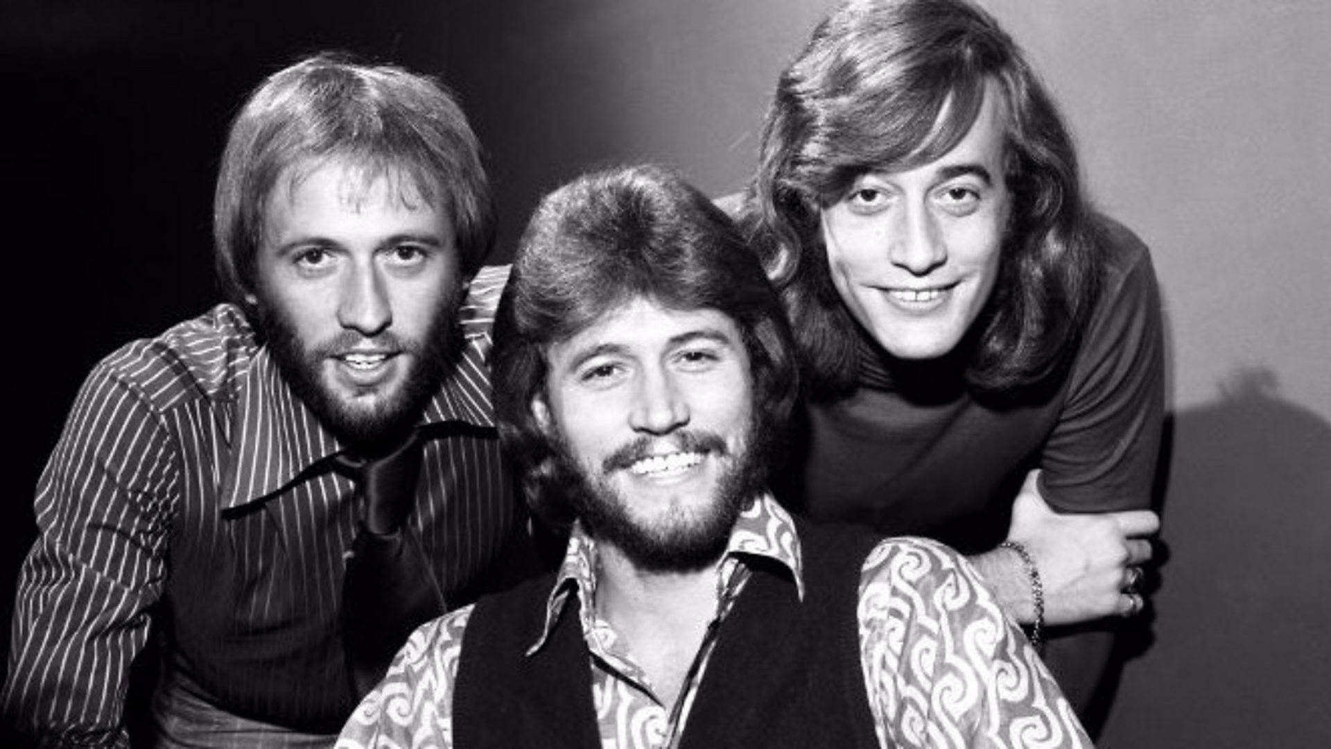 Bee Gees 三兄弟復古肖像 壁紙 — 三位長髮、身穿圖案襯衫的微笑男士，在經典黑白工作室肖像中緊密擺姿勢。