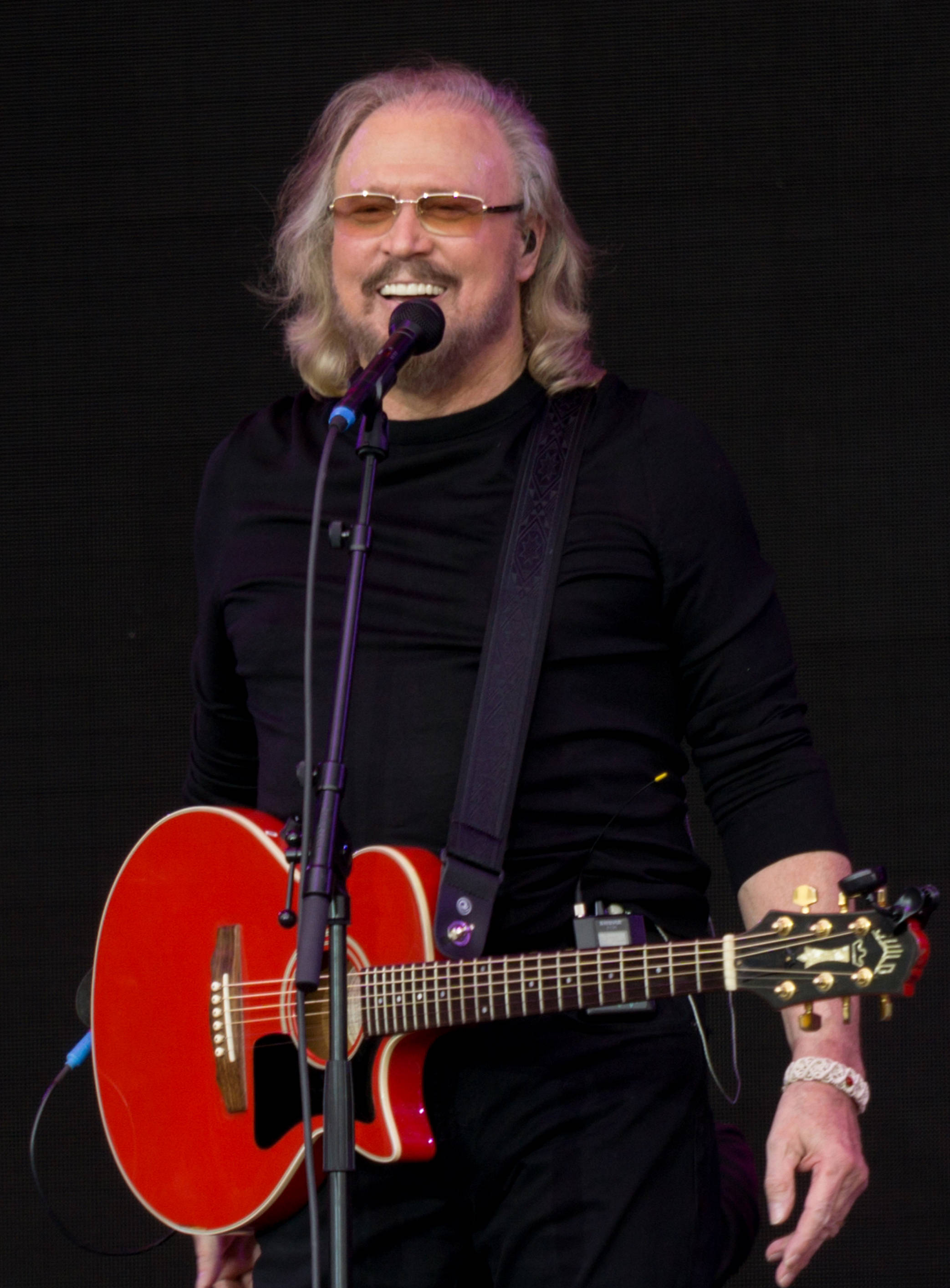 Bee Gees 成員巴里·吉布 2017 年格拉斯頓伯里音樂節演出 壁紙 — 一位微笑的長髮音樂家戴著太陽眼鏡，在明亮的燈光下於舞台上彈奏紅色原聲結他。