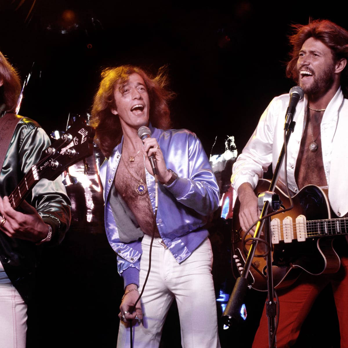Bee Gees 流行樂團現場演出 壁紙 — 三位長髮搖滾音樂家在明亮的燈光下，手持結他和咪高峰，在舞台上充滿活力地表演。