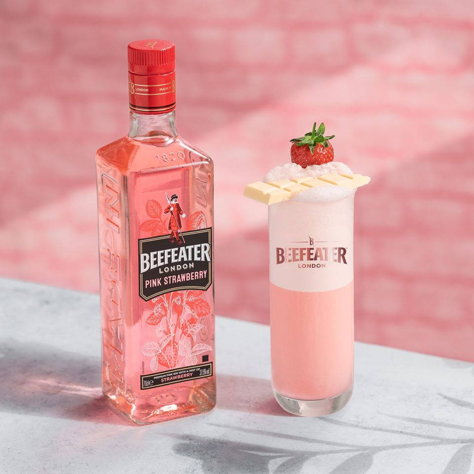 Beefeater 草莓白巧克力口味 壁紙 — 一瓶Beefeater Pink Strawberry氈酒放在一杯粉紅色雞尾酒旁邊，雞尾酒上放有白朱古力和草莓，背景是柔和的粉紅色磚牆。
