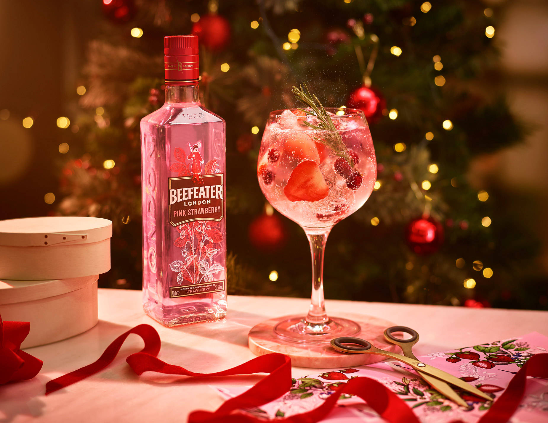 Beefeater聖誕樹主題 壁紙 — 一款節日粉紅氈酒雞尾酒，以草莓和迷迭香裝飾，放在酒瓶旁邊，背景是閃爍的聖誕樹。