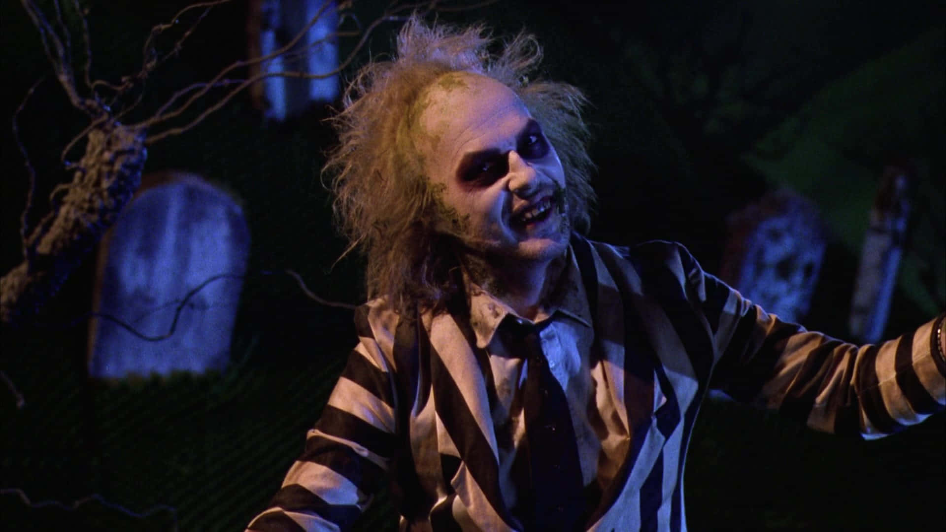 嘩鬼家族 桌布 壁紙 — Beetlejuice 穿著其標誌性條紋西裝，在陰森藍光下的黑暗墓地中狂笑。