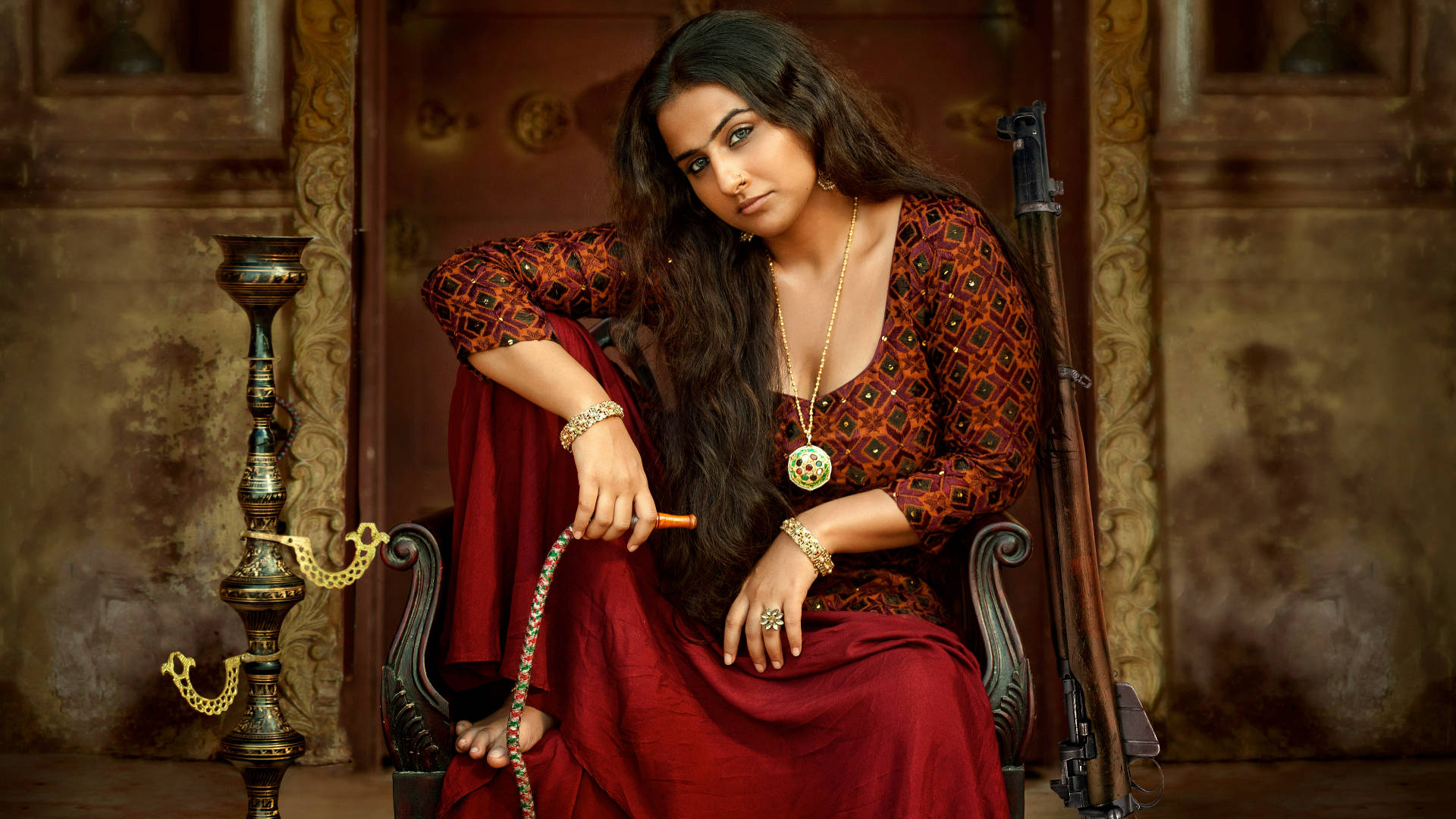 薇迪亞·巴蘭《Begum Jaan》 壁紙 — 一位身穿華麗紅色服飾的女性，端莊地坐在椅子上，手持水煙壺，身旁靠著一支步槍，身處裝飾華麗的房間。
