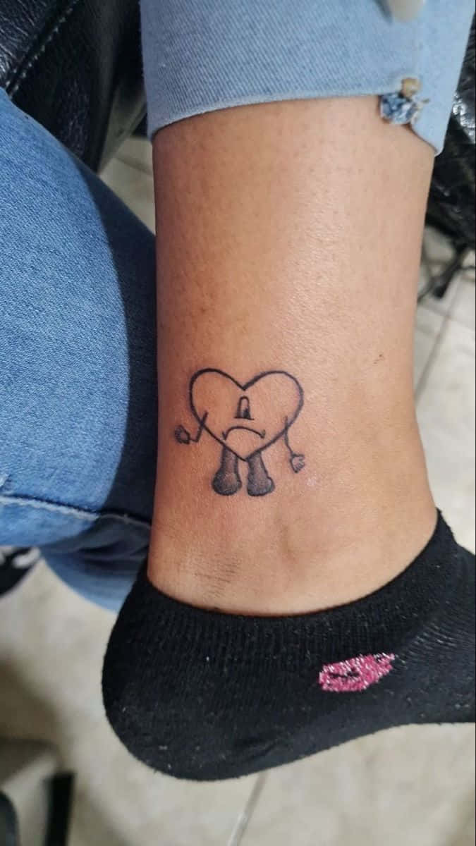 Bad Bunny Heart 桌布 壁紙 — 一個細小的黑色墨水紋身，圖案是一個悲傷的卡通心形，有手有腳，位於一個人的腳踝上。
