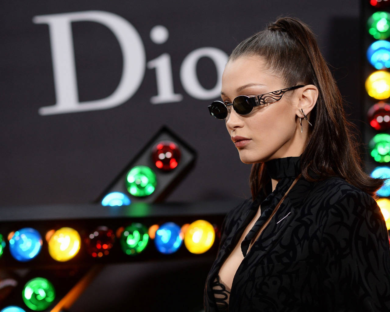 Bella Hadid 出席 Christian Dior 秀 壁紙 — 一位身穿黑色圖案服裝和華麗太陽眼鏡的女性，在閃爍的多彩燈光展示前擺姿勢，背景是Dior標誌。