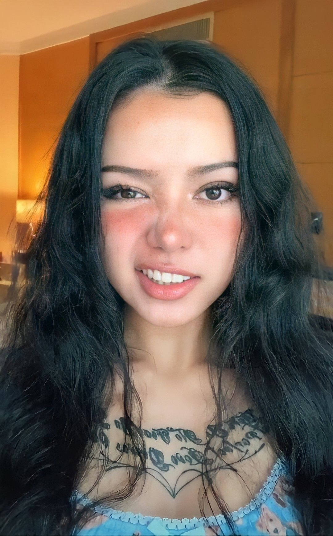 Bella Poarch 可愛近照 壁紙 — 一位擁有長長黑髮和胸口紋身的年輕女性，在溫暖的室內環境中露出溫暖的笑容。