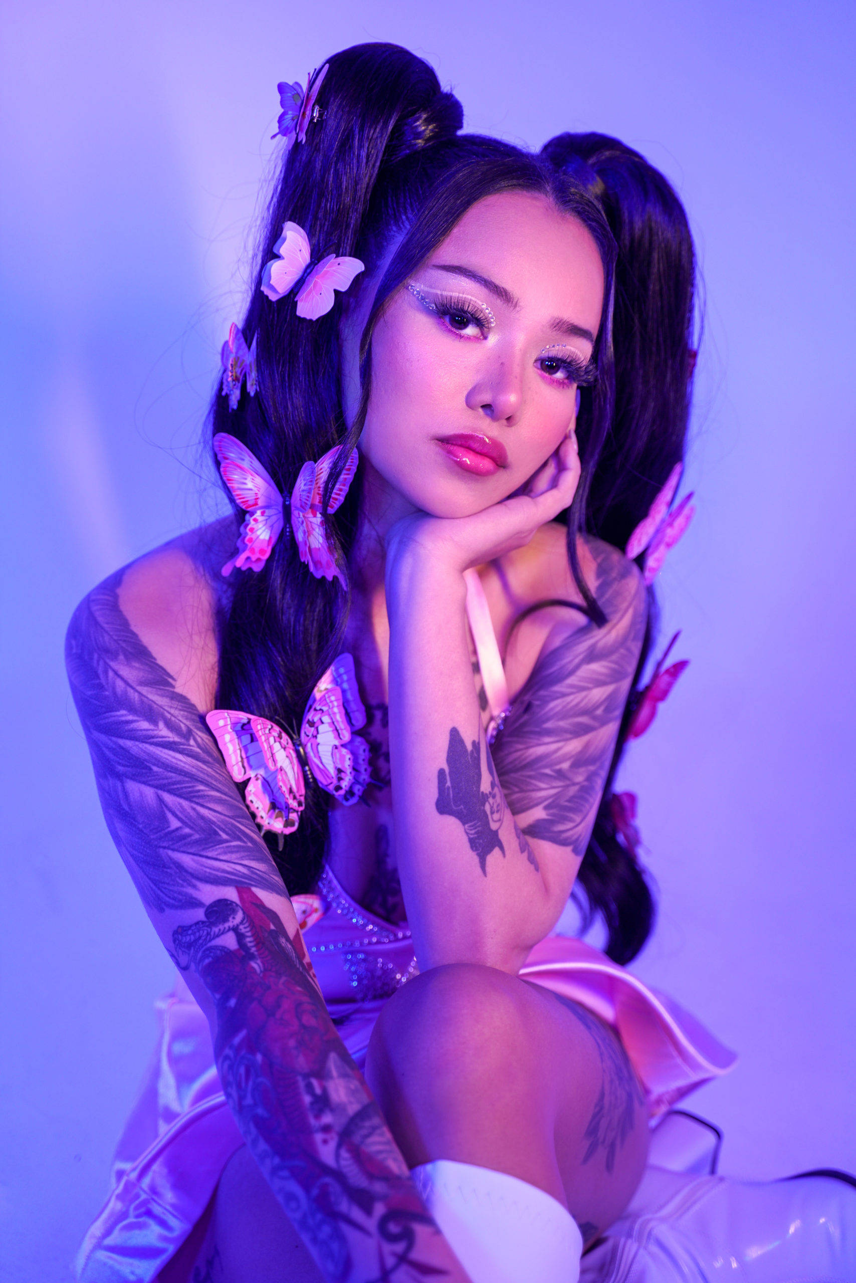 Bella Poarch 萌系蝴蝶髮型寫真 壁紙 — 一位戴著蝴蝶髮夾和紋身的女性在工作室的紫色燈光下擺姿勢。