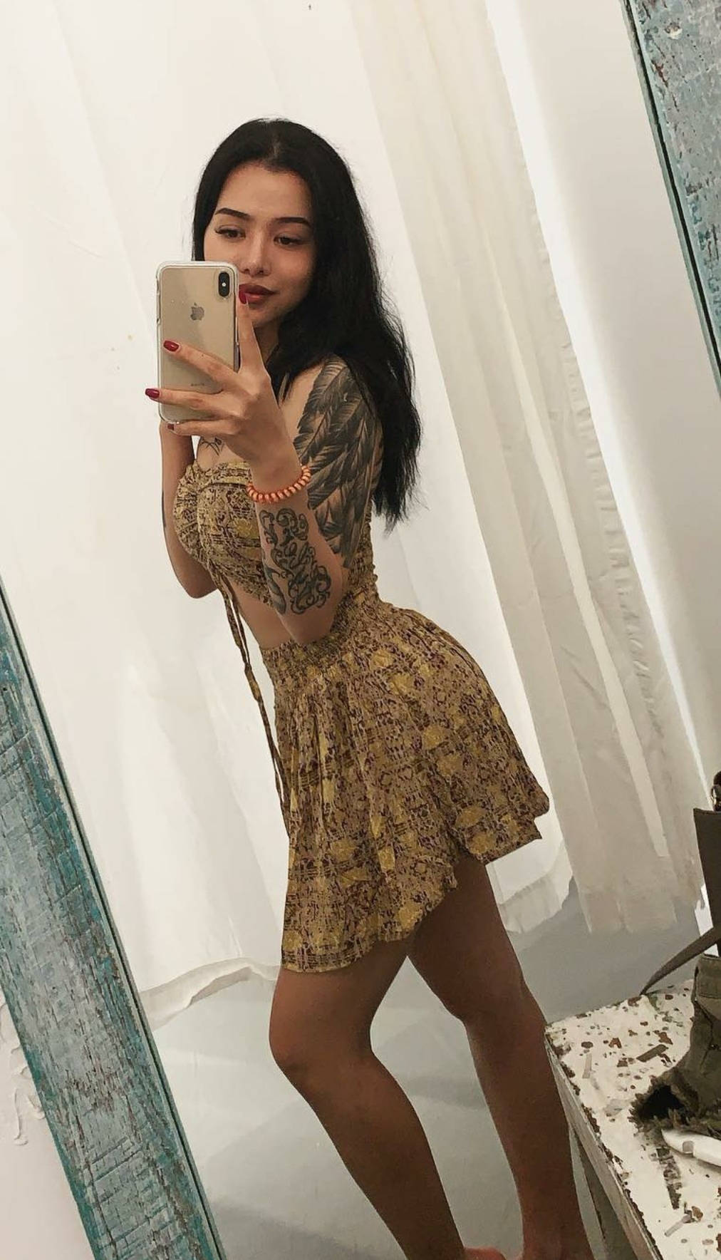 Bella Poarch 鏡頭自拍照 壁紙 — 一位深色頭髮、有紋身的女性，穿著圖案連衣裙，在白色窗簾背景下對著鏡子自拍。