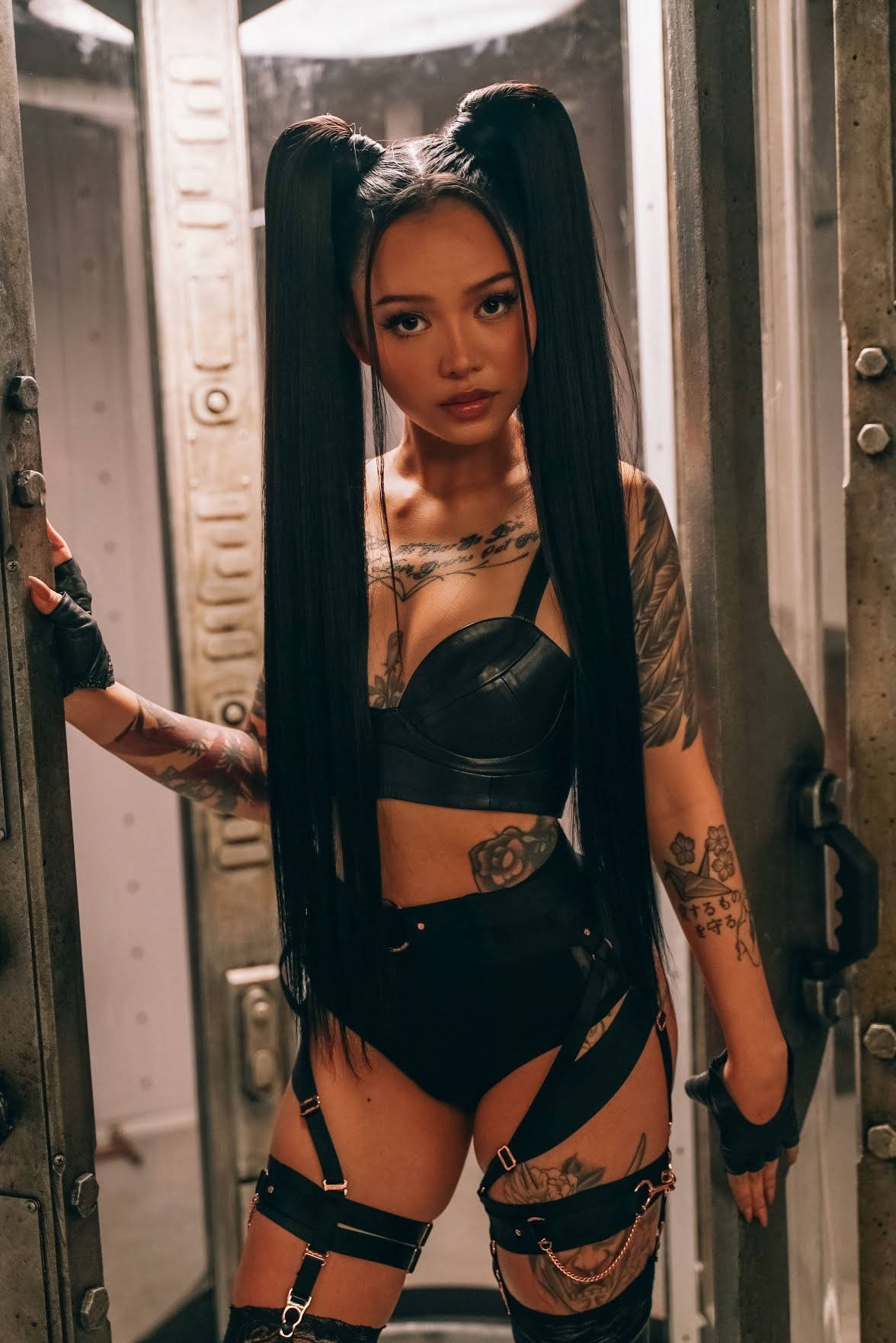 Bella Poarch熱門造型 壁紙 — 一位紋身長辮女性自信地站在金屬門口，身穿黑色皮革和束帶。