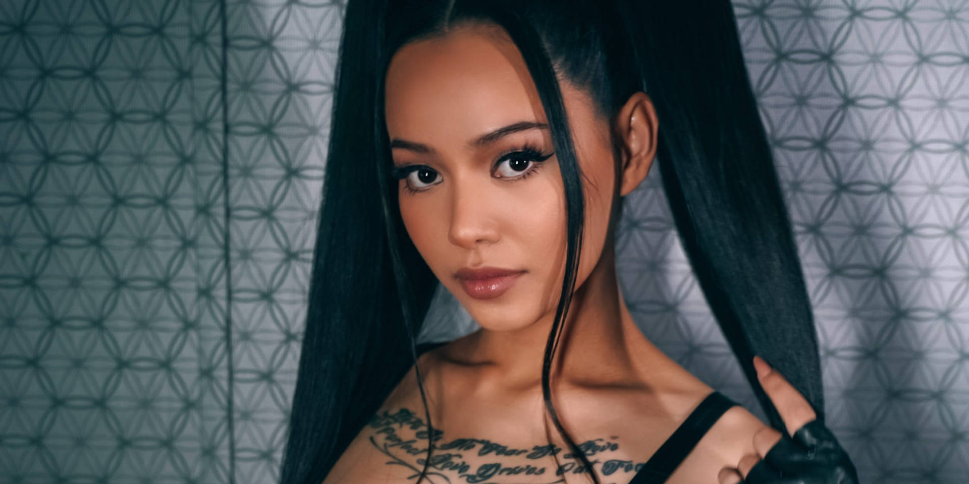 Bella Poarch雙馬尾特寫 壁紙 — 一位擁有長黑髮和紋身的女性，在幾何圖案牆壁前直視鏡頭。