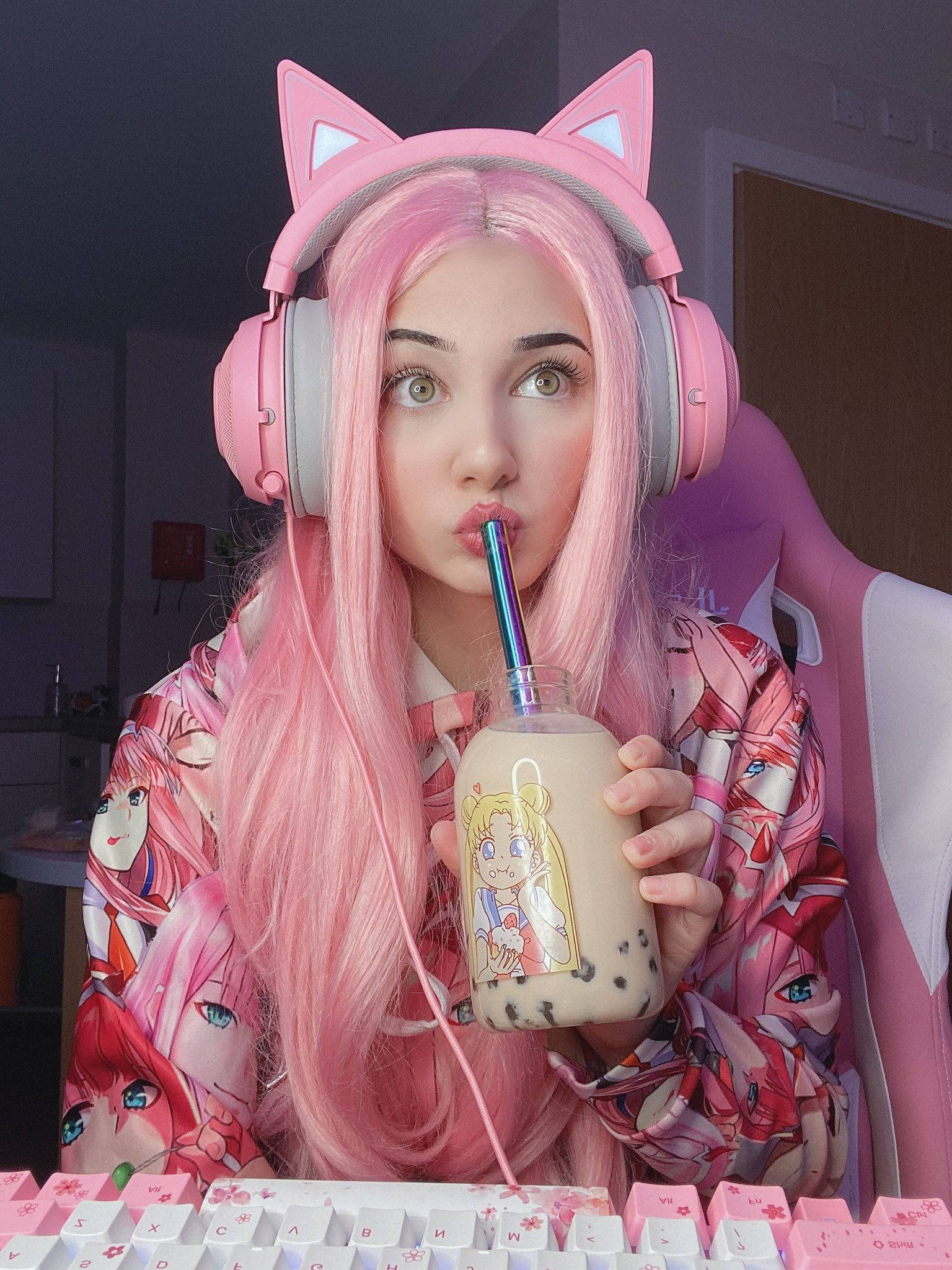 Belle Delphine 喝奶茶 壁紙 — 一位年輕女性，粉紅色頭髮，戴著貓耳耳機，坐在粉紅色鍵盤旁喝珍珠奶茶。