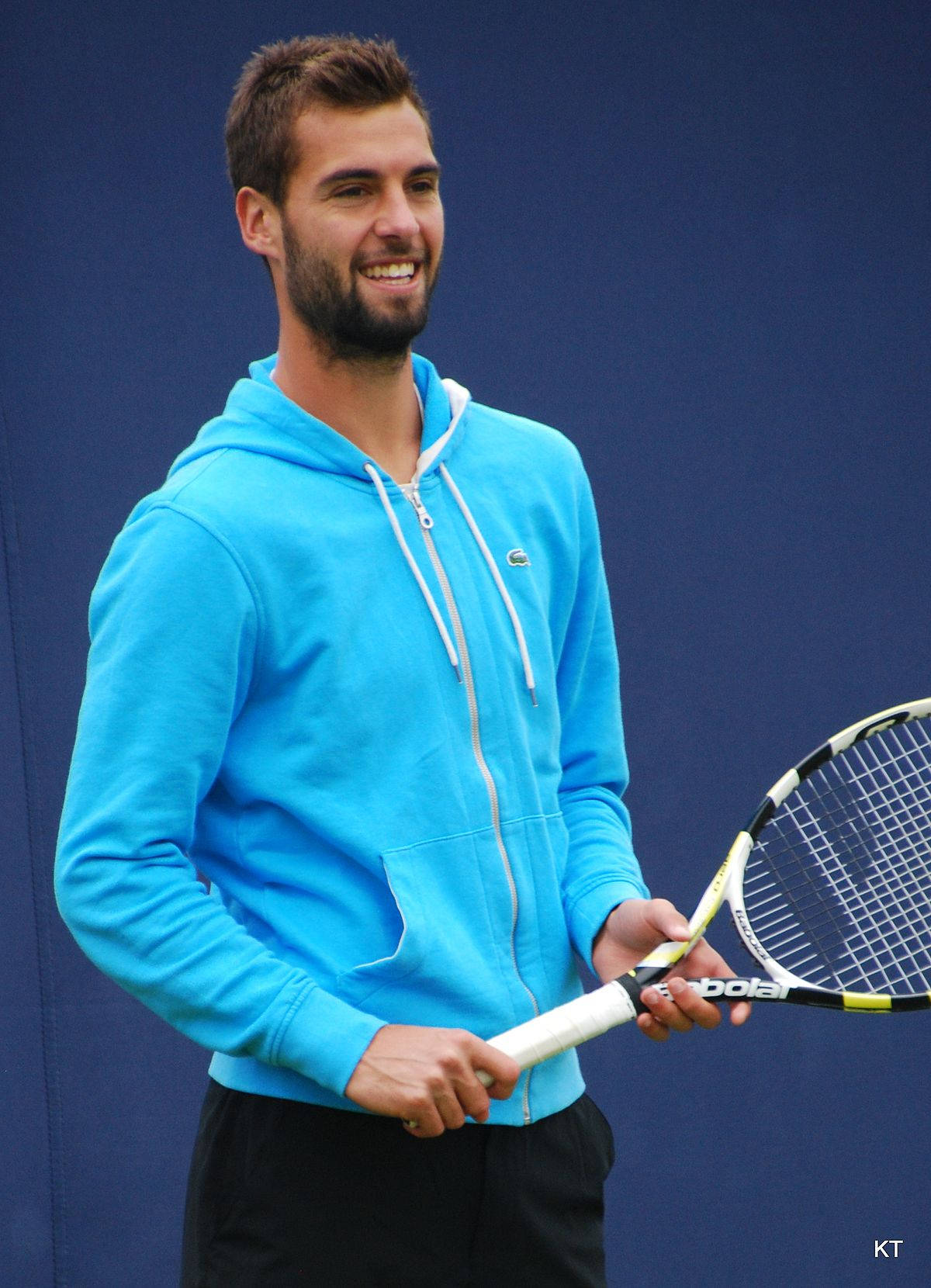 Benoit Paire 微笑握拍 壁紙 — 一個微笑的男人穿著亮藍色連帽衫，在純藍色背景前拿著網球拍。