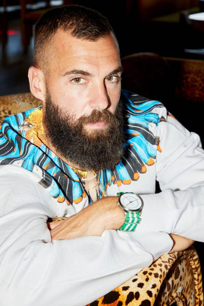 Benoit Paire 當模特兒 壁紙 — 一位留鬍鬚的男子穿著彩色圖案連帽衫和綠色手錶，在昏暗的房間裡將手臂擱在豹紋椅子上。