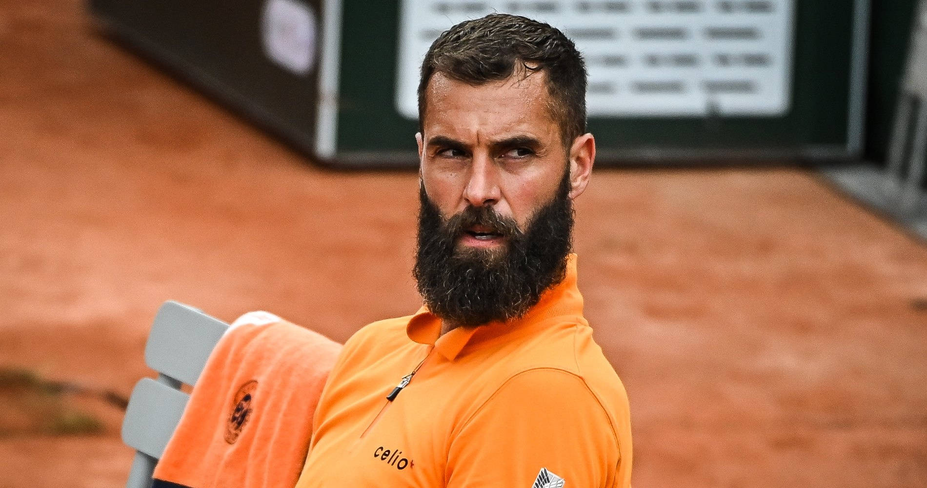 Benoit Paire 瞪眼凝視 壁紙 — 一位留鬍鬚的網球運動員穿著橙色Polo衫，專注地坐在紅土球場的長凳上。
