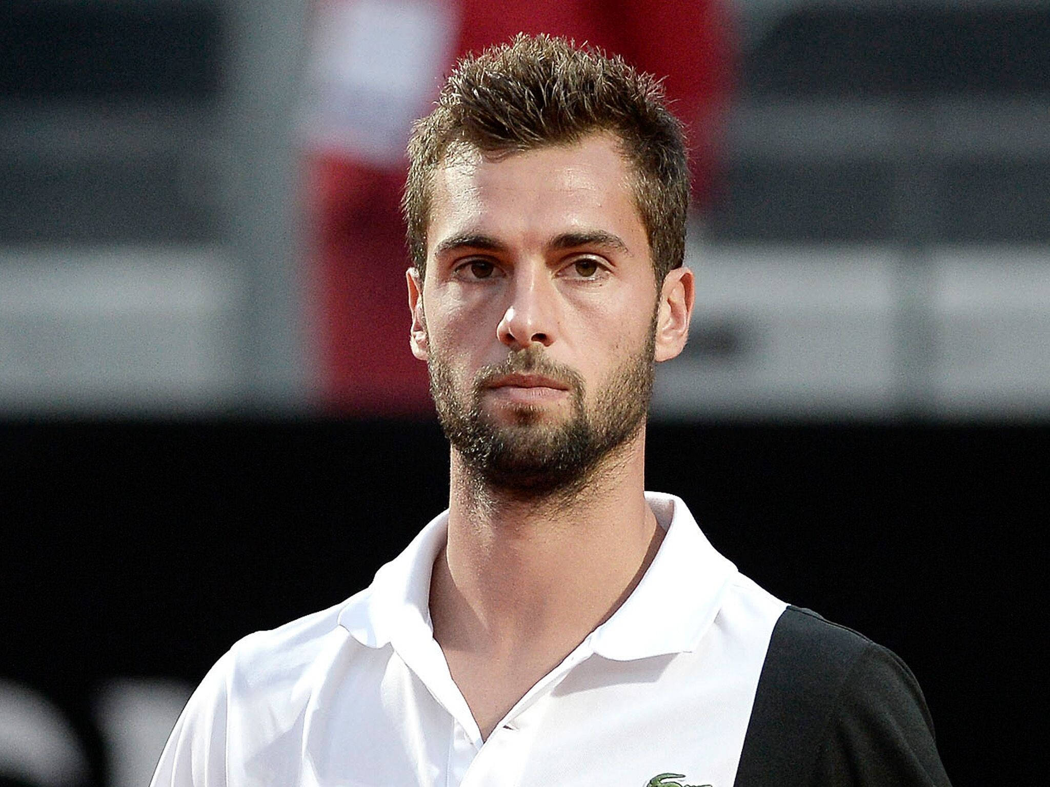 Benoit Paire 細節特寫嚴肅表情 壁紙 — 一位留鬍鬚、短髮的專注男網球運動員，穿著白色和黑色Polo衫，背景是模糊的體育場。
