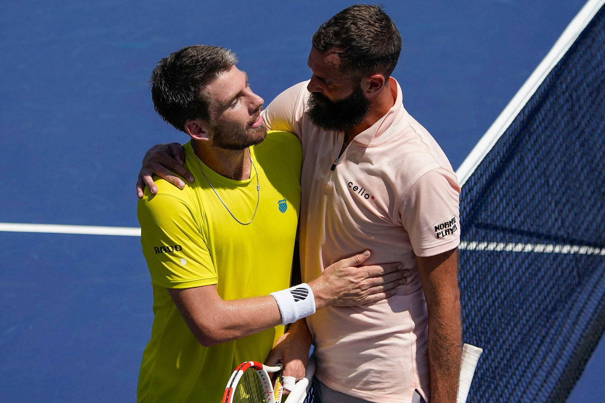Benoit Paire 與 Cameron Norrie 賽後擁抱 壁紙 — 兩名男網球運動員在明亮的日光下，在充滿活力的藍色球場上熱情擁抱。