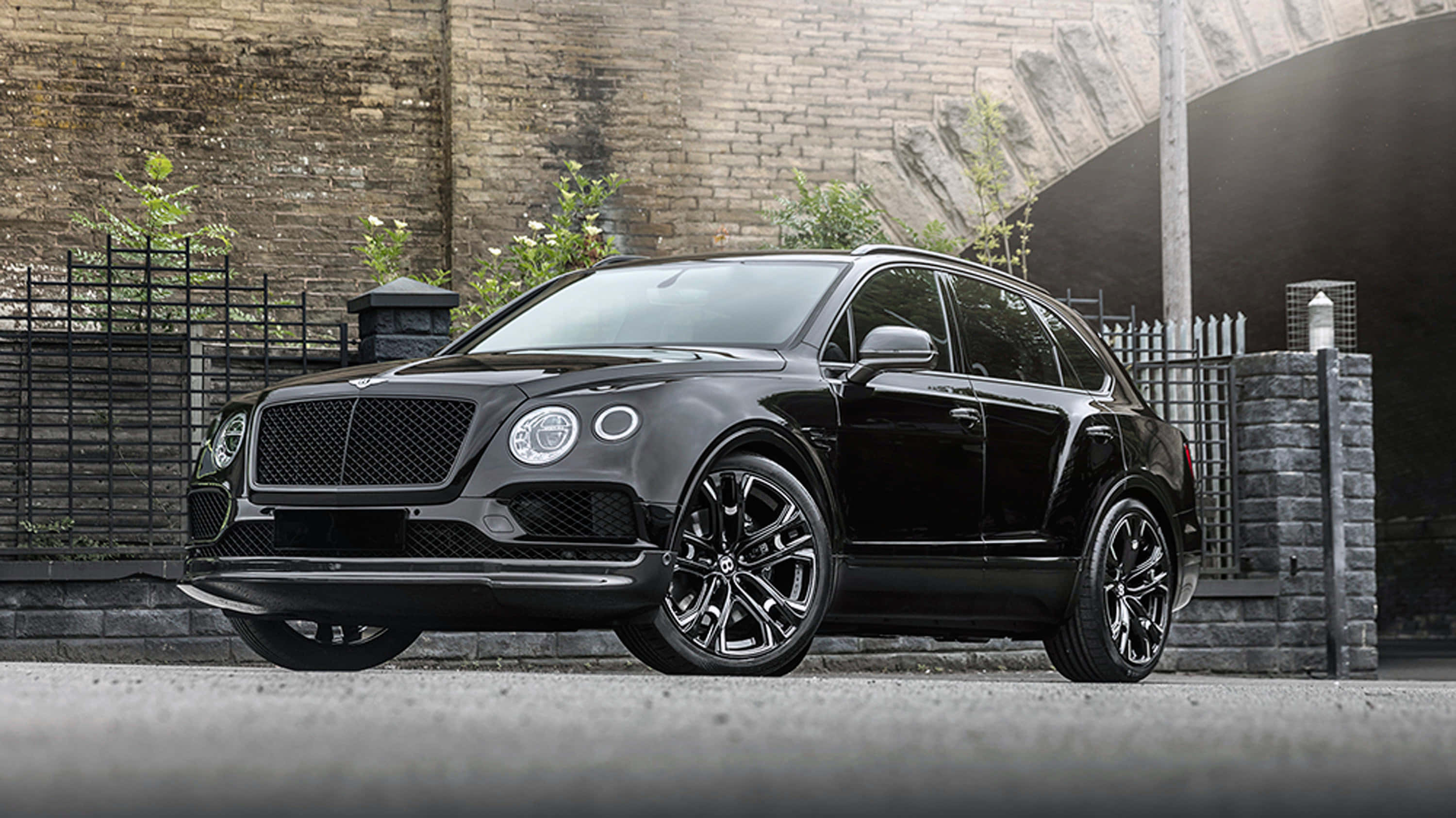豪華賓利Bentayga SUV駛過開放道路 壁紙 — 一輛時尚的黑色賓利Bentayga SUV停在風化的磚牆和金屬圍欄前。
