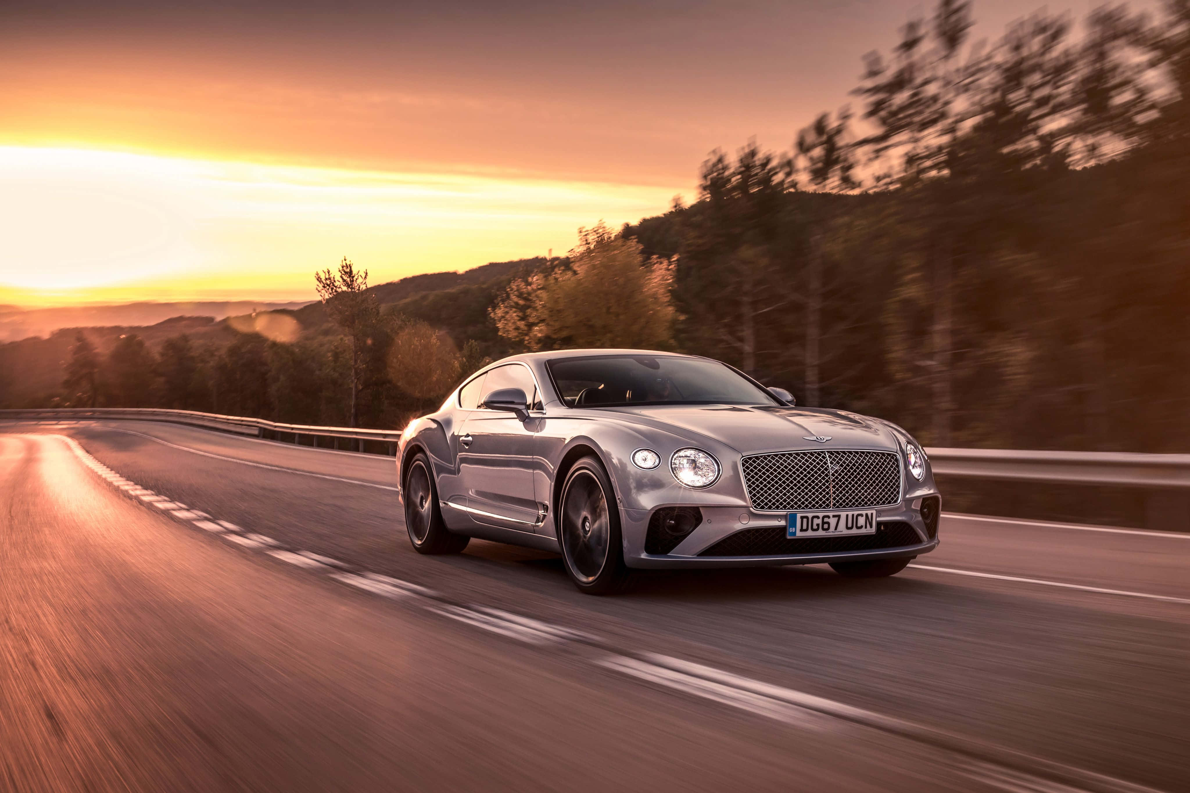 夢幻奢華座駕：Bentley 壁紙 — 一輛銀色賓利 Continental GT 在日落時分沿著蜿蜒的山路疾馳，金色光芒照亮了整個場景。