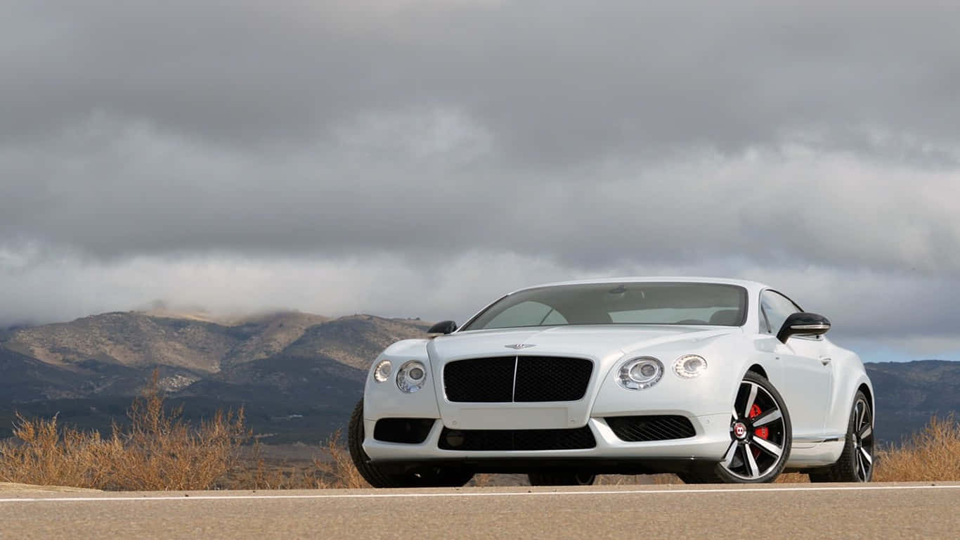 非凡氣質 — Bentley 壁紙 — 一架流線型白色賓利 Continental GT 雙門跑車停泊在路邊，背景是崎嶇的山脈和陰天。